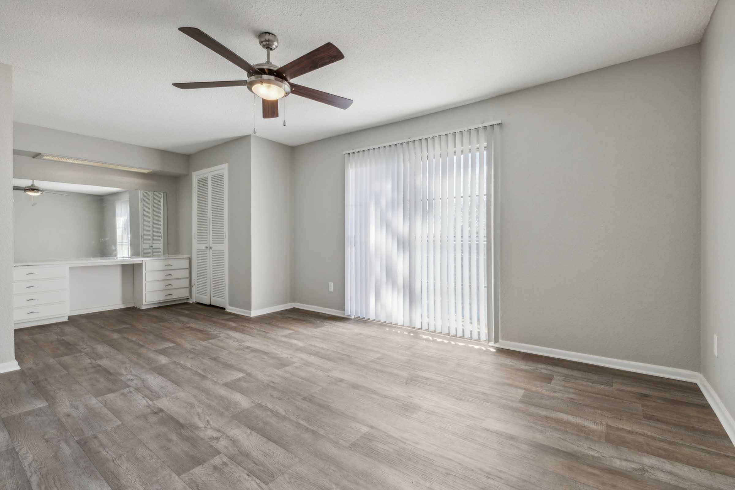 A spacious room with light brown wooden floor, ceiling fan, and large window with white blinds letting in natural light. There is a white vanity with drawers against one wall, and a small closet. The walls are painted a light gray, creating a clean and modern look.