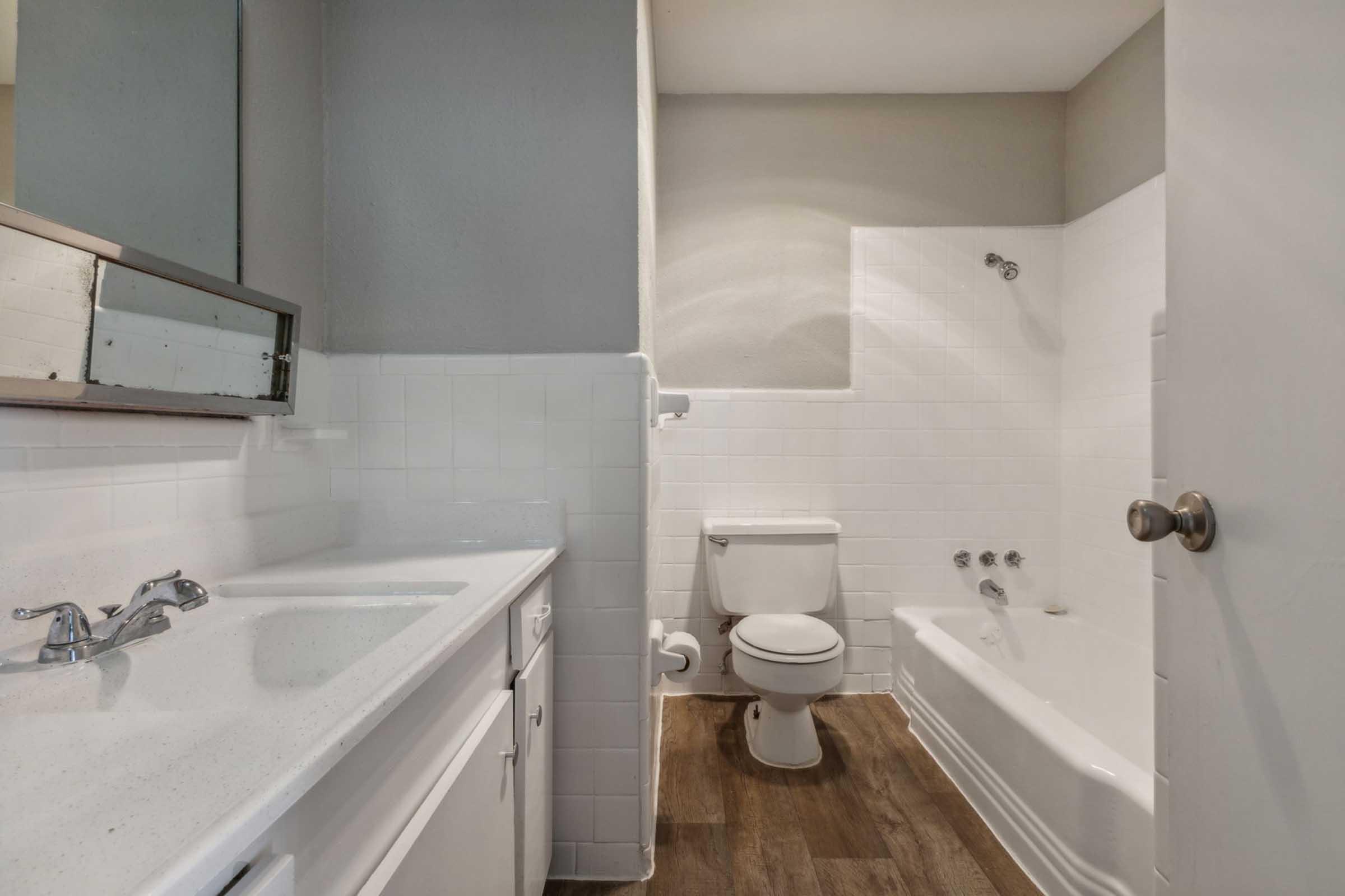 A clean and modern bathroom featuring a white tiled bathtub with a shower attachment, a toilet, and a sink with a minimalist vanity. The walls are painted in neutral tones, and there is a large mirror above the sink, creating a bright and spacious feel.