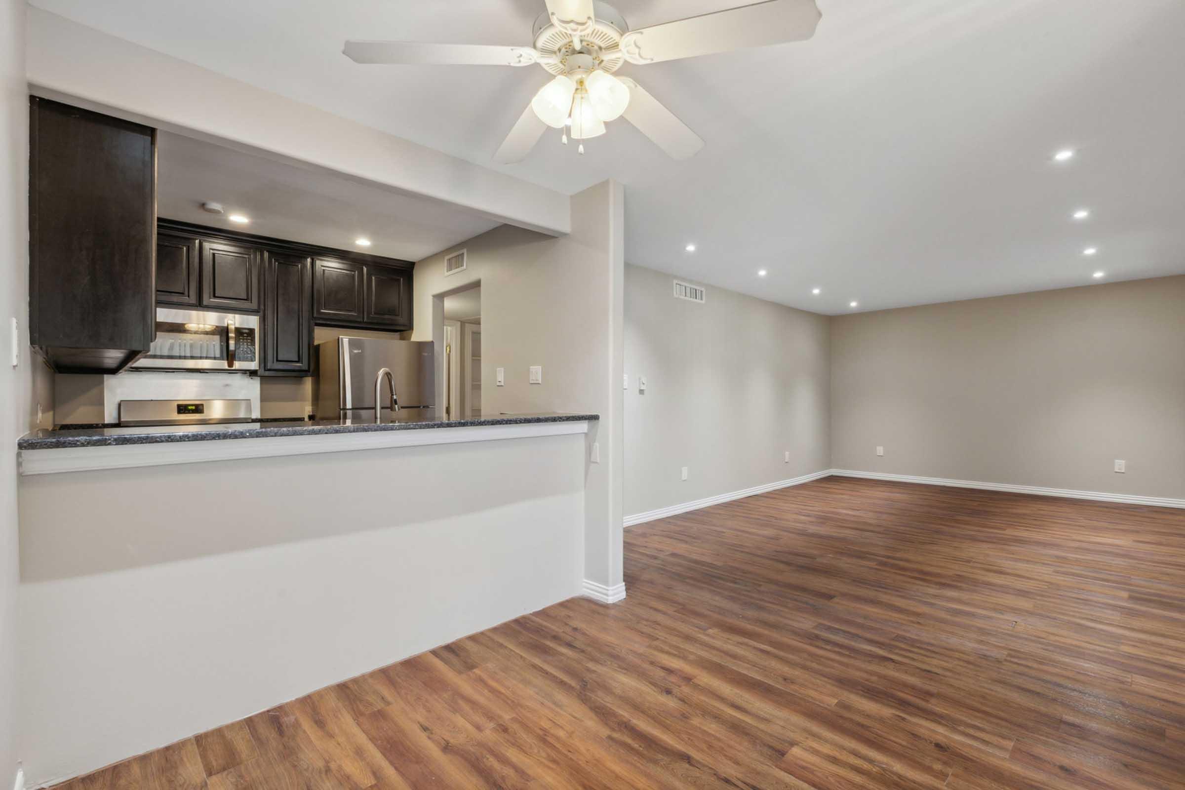 Interior view of a spacious living area featuring hardwood floors, a ceiling fan, and a modern kitchen with dark cabinetry. The room is well-lit with recessed lighting and has neutral-colored walls, creating a warm and inviting atmosphere.