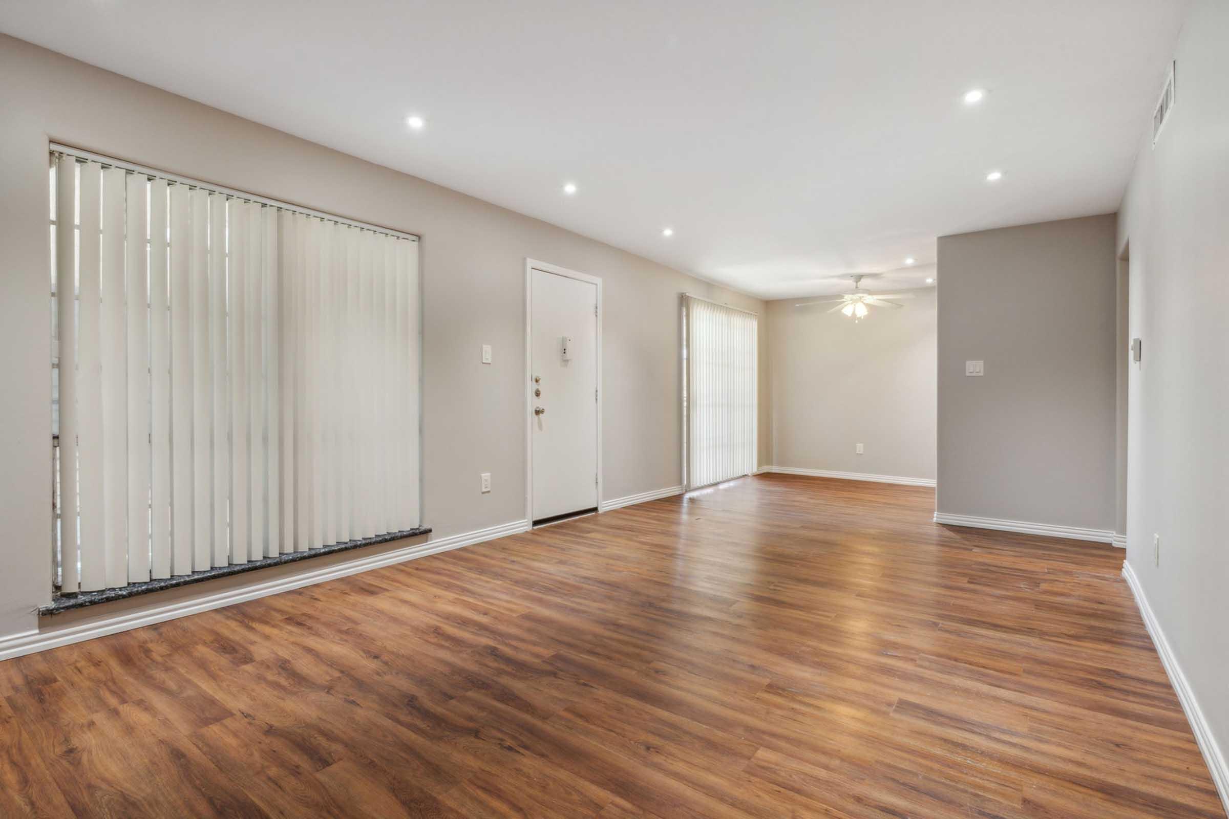 A spacious and empty living room with hardwood floors. There are two large windows with vertical blinds, one door leading outside, and a ceiling fan. The walls are painted in a neutral color, creating a bright and airy ambiance.
