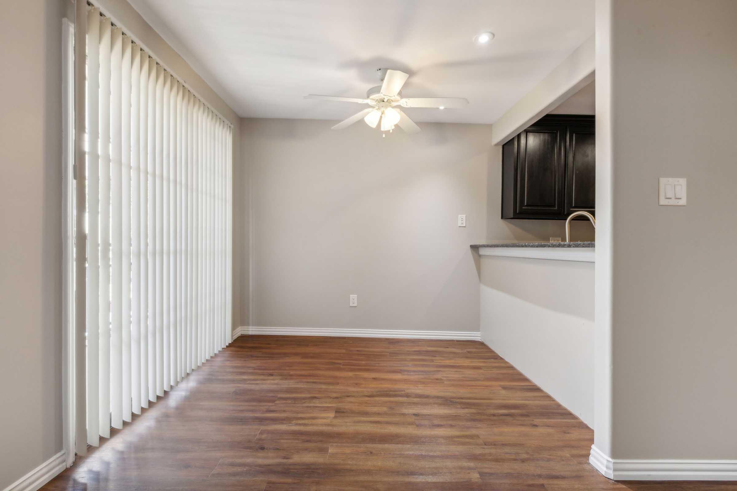 A spacious room with hardwood flooring, featuring a ceiling fan and large sliding glass doors with vertical blinds. The walls are painted in a light color, and a small kitchen area with dark cabinetry is visible in the background. Natural light fills the space, creating a bright atmosphere.