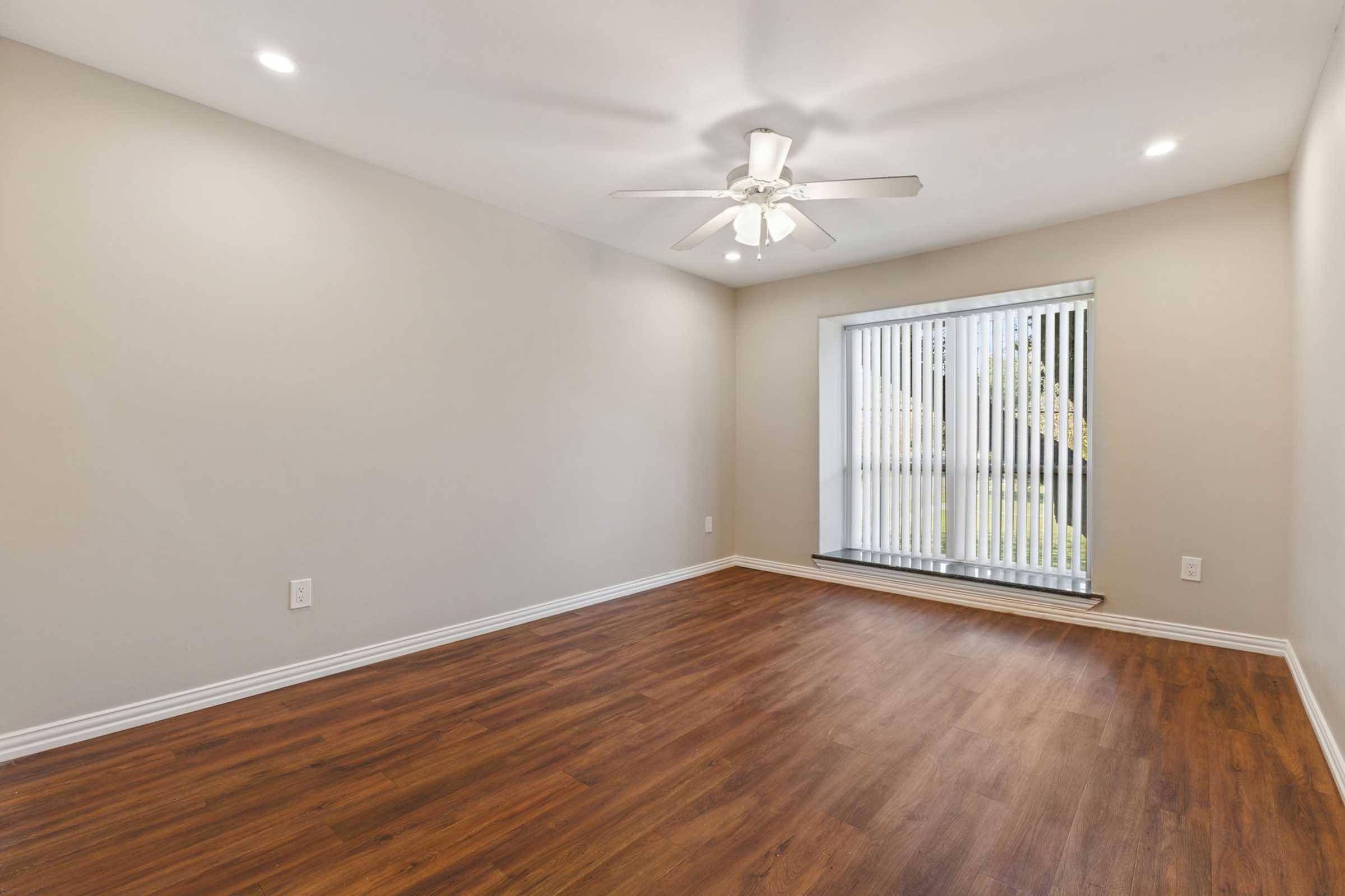 Empty room with light brown hardwood flooring, a ceiling fan, and a window featuring vertical blinds. The walls are painted a light neutral color, and there are recessed lights in the ceiling. The overall atmosphere is bright and spacious.