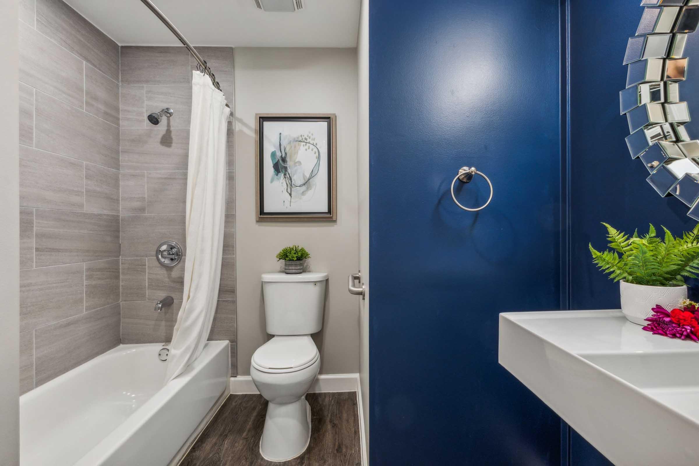 A modern bathroom featuring a bathtub with a shower, a toilet, and a small decorative plant. The walls are tiled in light gray, with a dark blue accent wall. A round, mirrored bathroom mirror hangs above a sleek white sink, complemented by colorful flowers on the counter.