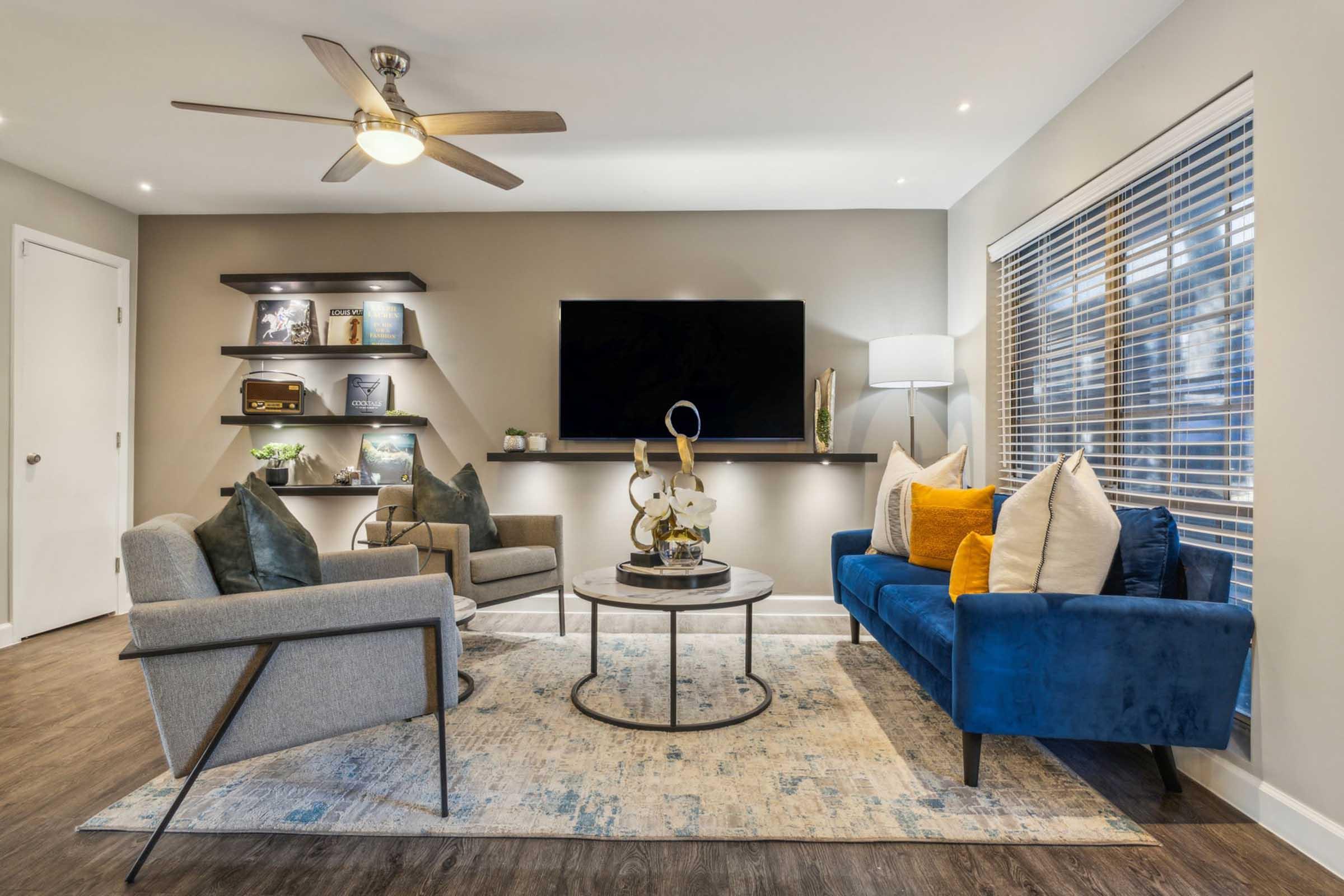 A modern living room featuring a blue sofa and gray armchairs, a round coffee table with decorative items, a flat-screen TV mounted on the wall, and stylish shelves displaying decor. Large windows with blinds allow natural light, enhancing the cozy atmosphere. A ceiling fan adds a contemporary touch.