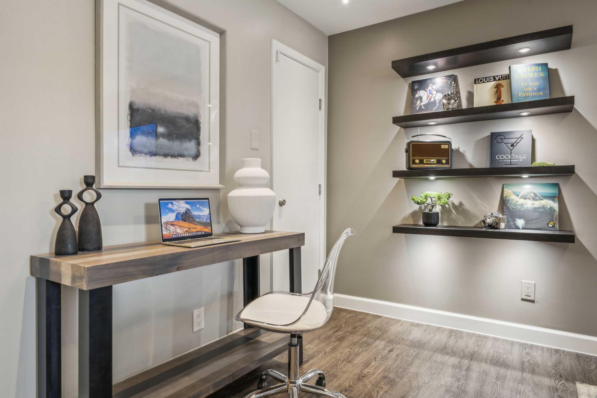 A modern workspace featuring a sleek desk with a laptop, a decorative vase, and an acrylic chair. The wall shelves are organized with books, a plant, and a record player, all set against a neutral-colored wall with framed artwork above the desk.
