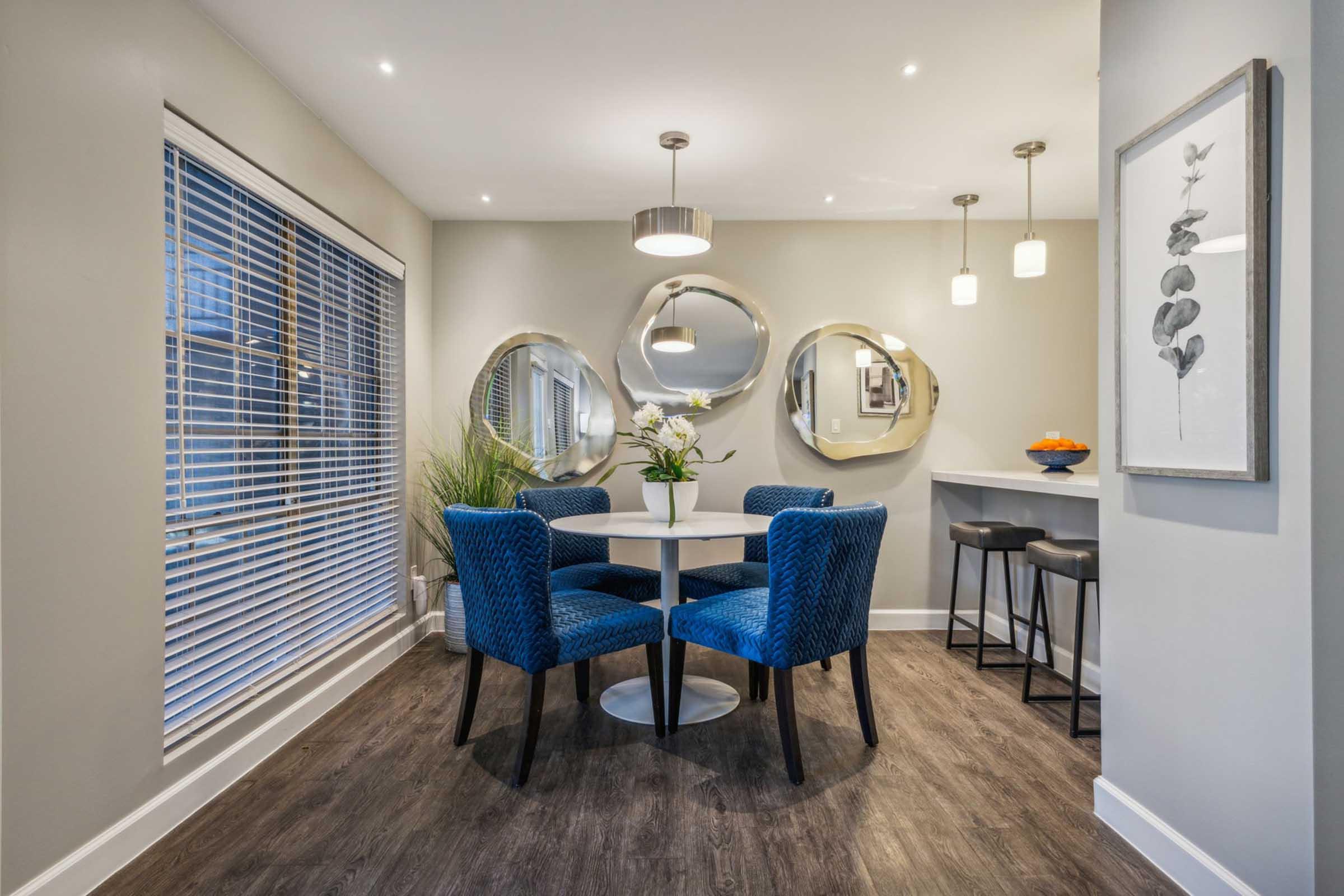A modern dining area featuring a round white table surrounded by four blue velvet chairs. The walls are light gray with large mirrors and a framed artwork. A bar area with two stools is visible in the background, and a window with blinds lets in natural light. A plant is in the corner, adding a touch of greenery.