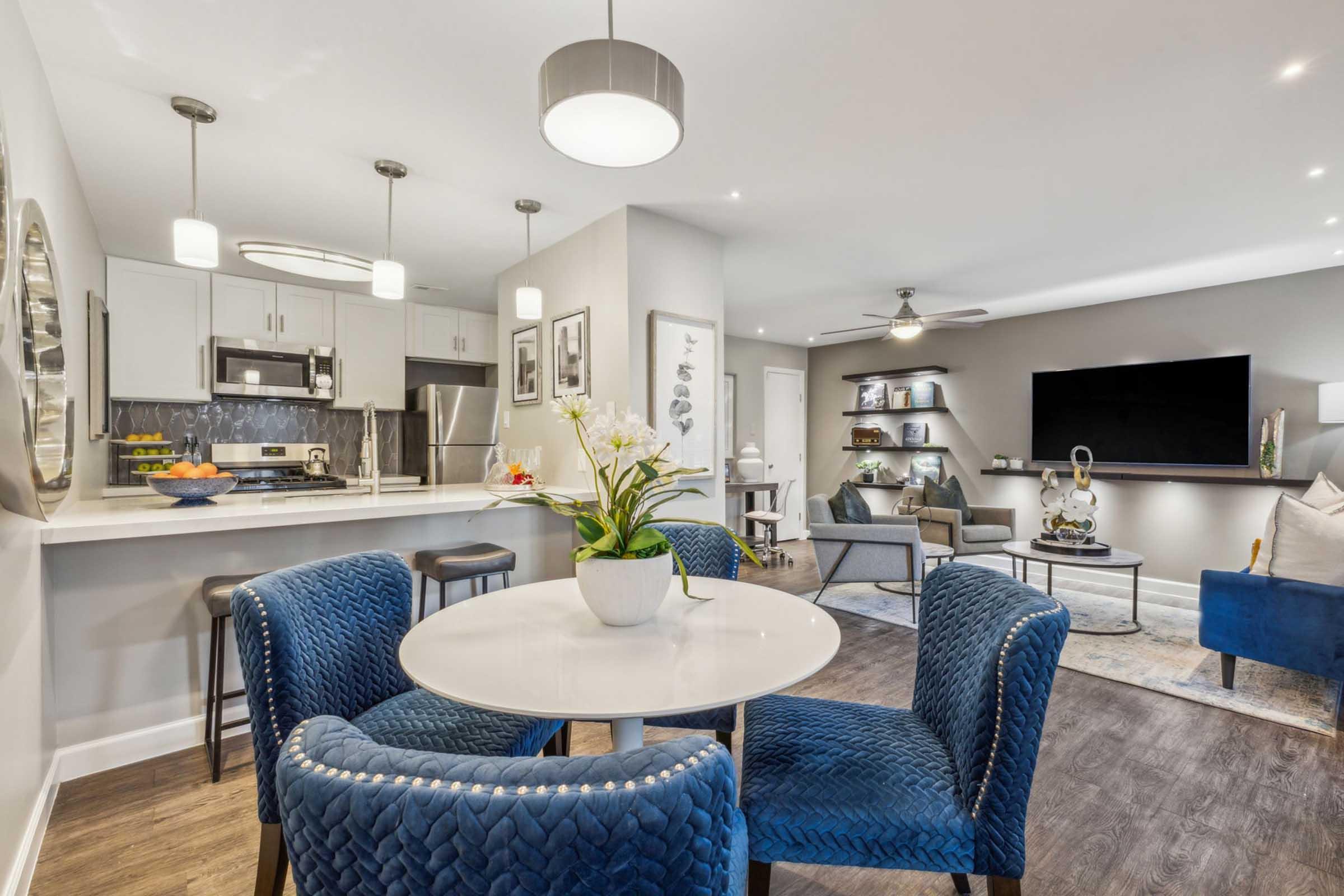 Modern living space featuring an open-plan layout. A round table with blue upholstered chairs is set in the center, surrounded by a kitchen with sleek cabinets and stainless steel appliances. A large TV is mounted on a wall in the living area, which includes stylish decor and a cozy armchair. A use of natural light enhances the atmosphere.