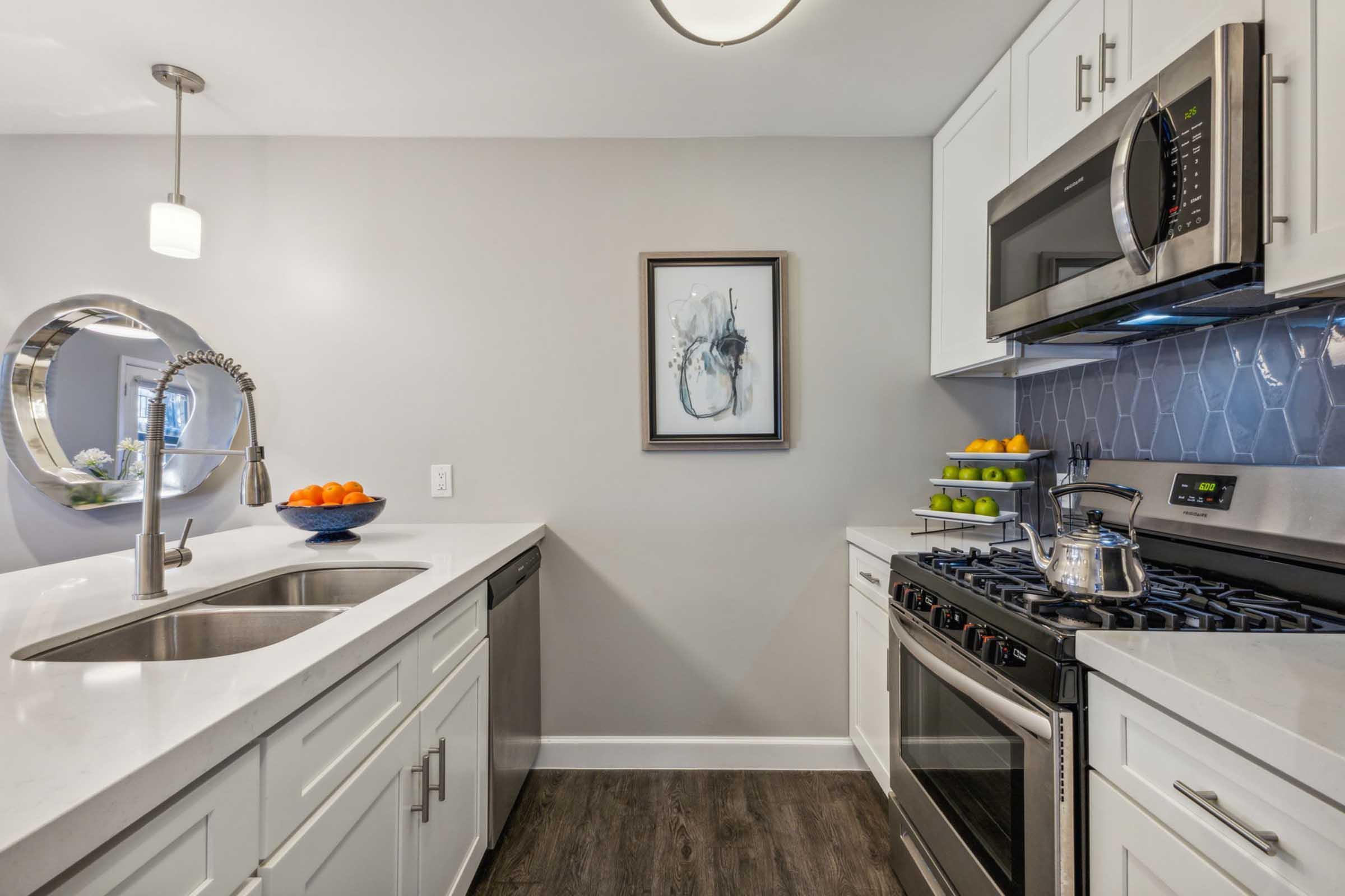 Modern kitchen featuring stainless steel appliances, including a gas range and microwave. A large sink with a sleek faucet sits next to a dishwasher. Decorative elements include a framed piece of art, a round mirror, and a bowl of oranges on the counter, complemented by a colorful display of fruit on a tiered stand.