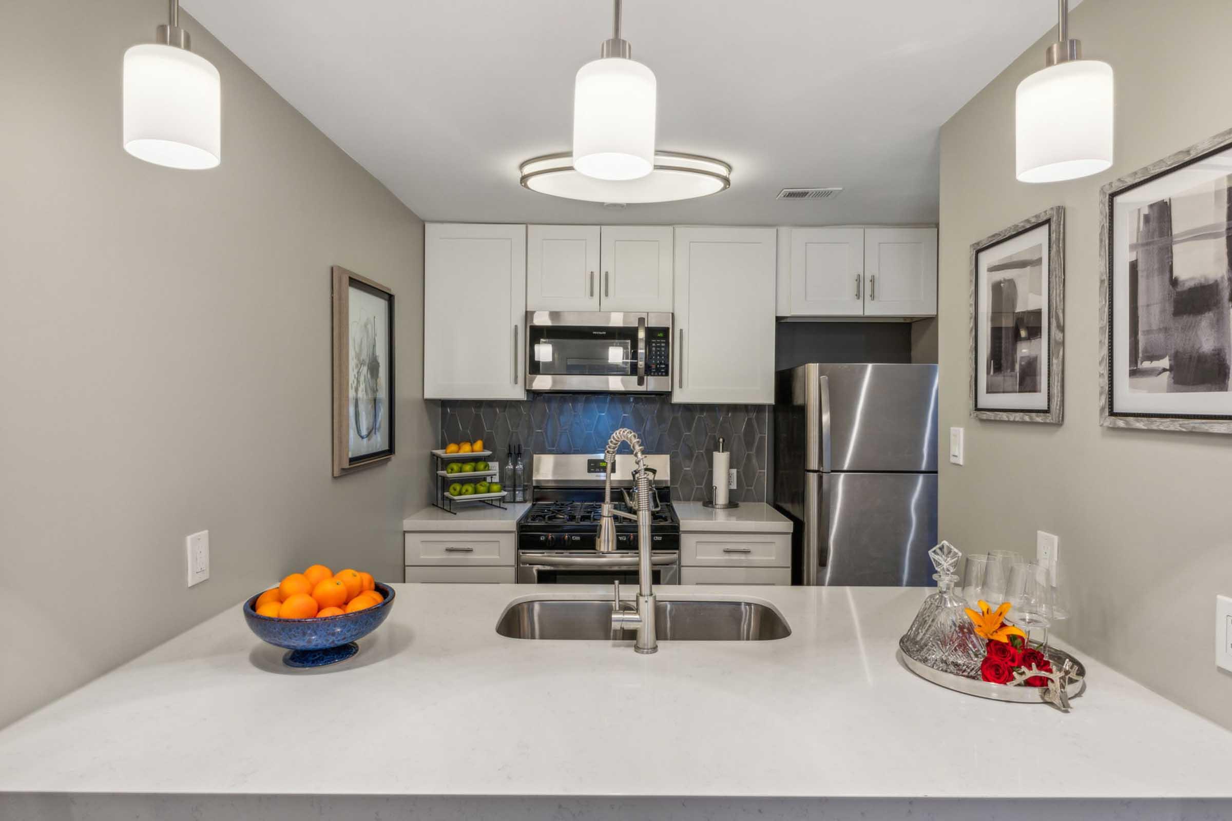 A modern kitchen featuring a marble countertop, stainless steel appliances, and white cabinetry. A bowl of oranges sits on the counter, and a decorative tray with flowers is visible. Pendant lights hang above the island, and framed artwork is displayed on the walls.