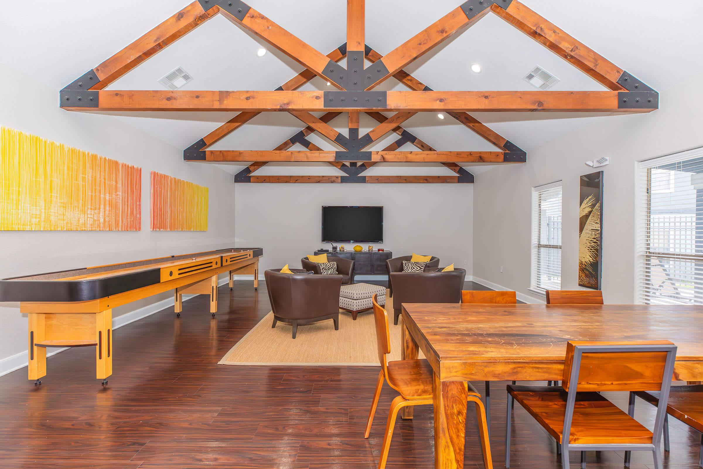 Spacious modern living area featuring a high, wooden-beamed ceiling, a large flat-screen TV, and comfortable seating with decorative cushions. The room includes a wooden dining table and chairs, a shuffleboard table, and colorful wall art in orange and yellow tones. Bright natural light streams through large windows.