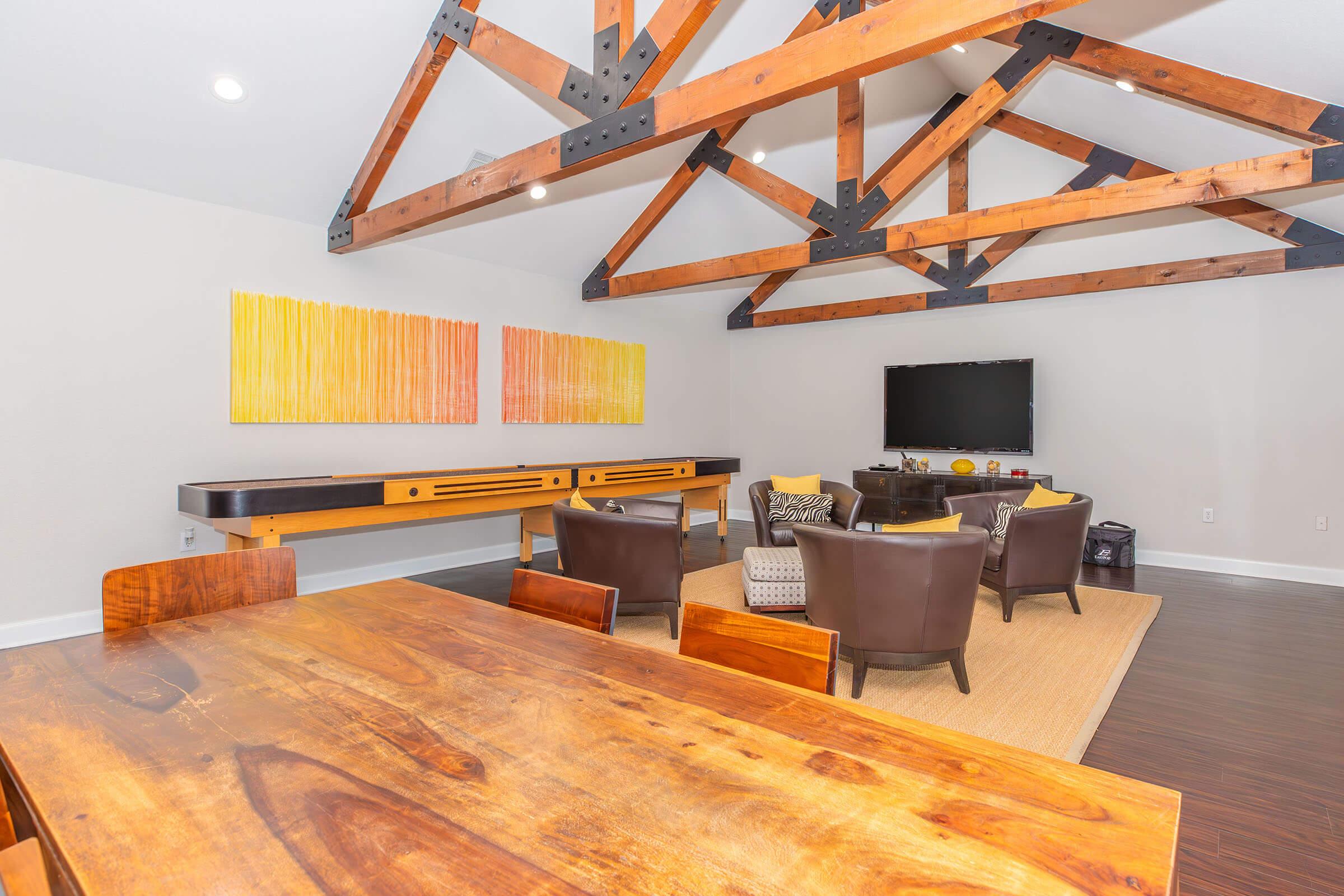 A spacious living room featuring high wooden beams, a large flat-screen TV, and modern furniture including an area rug, a coffee table, and stylish armchairs. Two abstract yellow and orange paintings adorn the wall. A wooden dining table is visible in the foreground, enhancing the warm, inviting atmosphere.