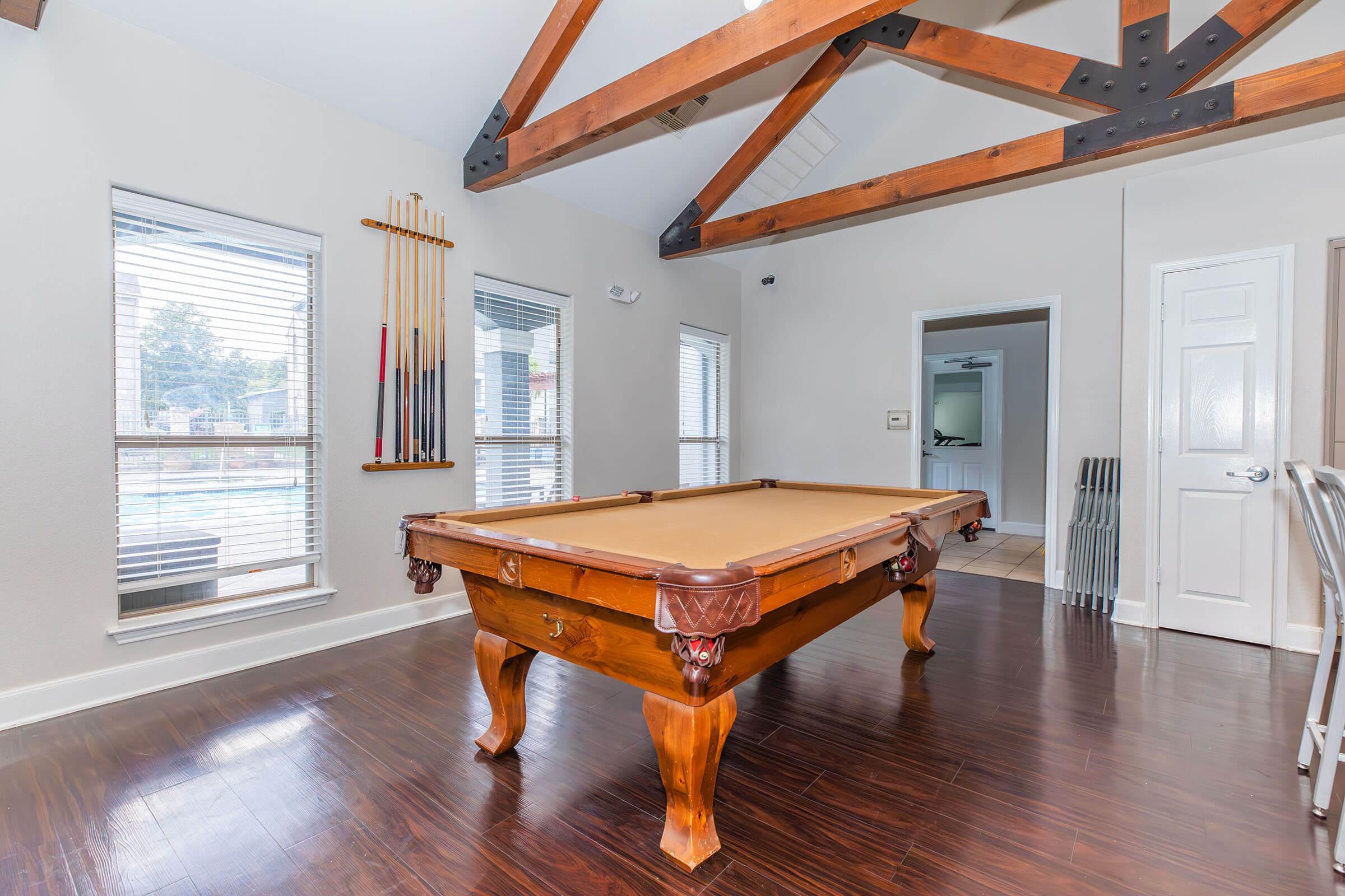 A spacious game room featuring a wooden pool table with intricate detailing, large windows allowing natural light, and a high ceiling with exposed wooden beams. Pool cues are neatly arranged on the wall, enhancing the recreational atmosphere of the room.