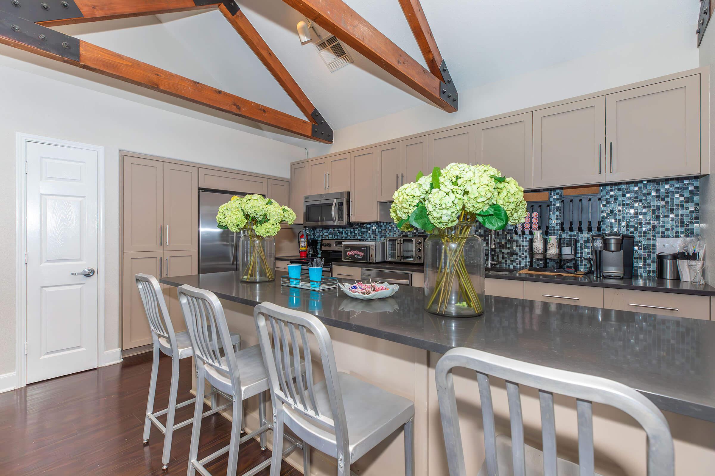 A modern kitchen featuring a large island with sleek gray countertops, three metal bar stools, and two vases filled with green flowers. The cabinets are a soft beige, and there are stainless steel appliances along with a decorative tile backsplash. Exposed wooden beams highlight the ceiling.