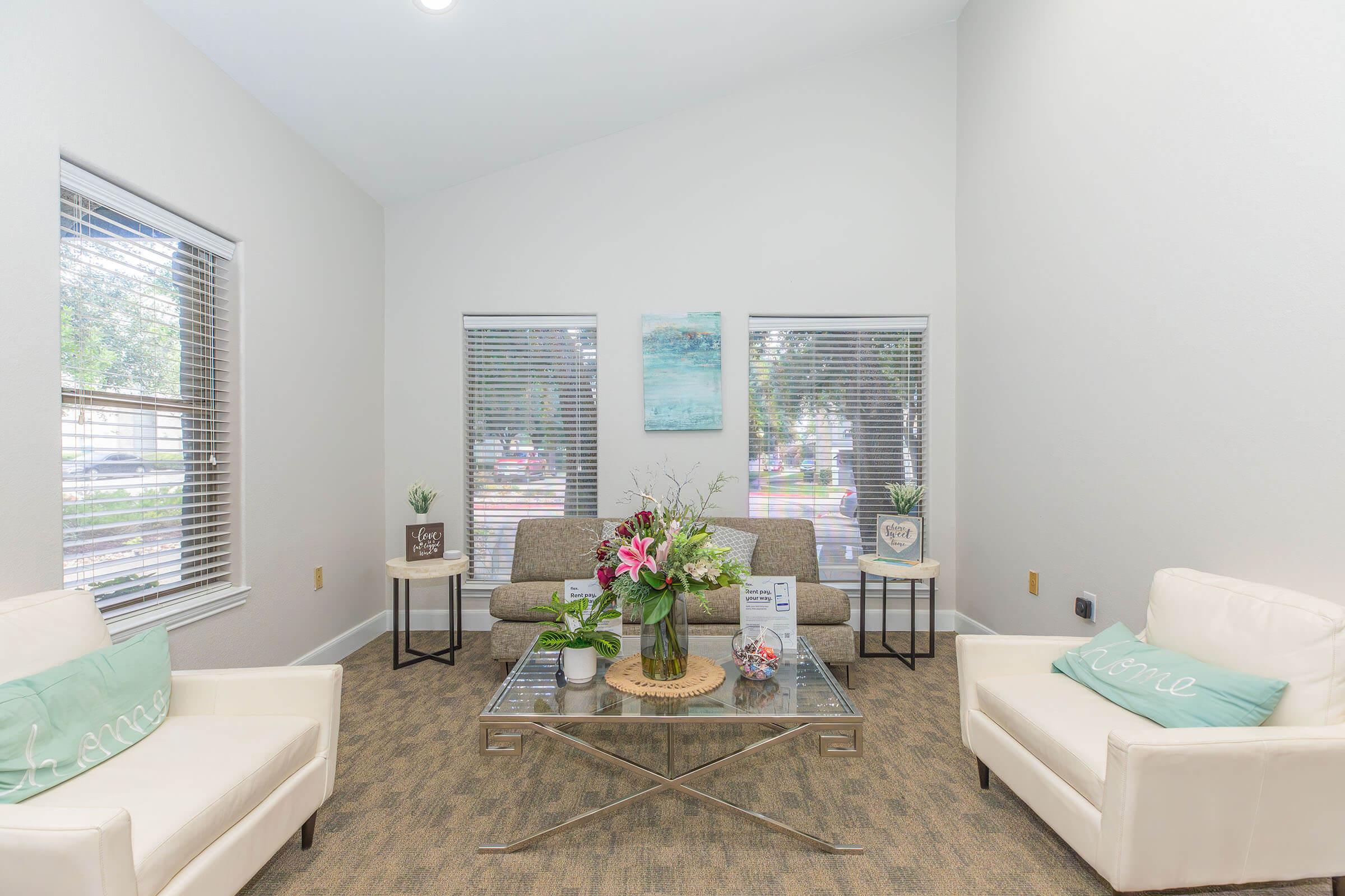 A cozy living room setup featuring a light gray sofa with decorative cushions, two white armchairs, and a glass coffee table adorned with flowers and decorative items. Natural light streams in through windows, complementing the neutral walls and carpeted floor. Decorative artwork hangs on the walls, inviting a warm ambiance.