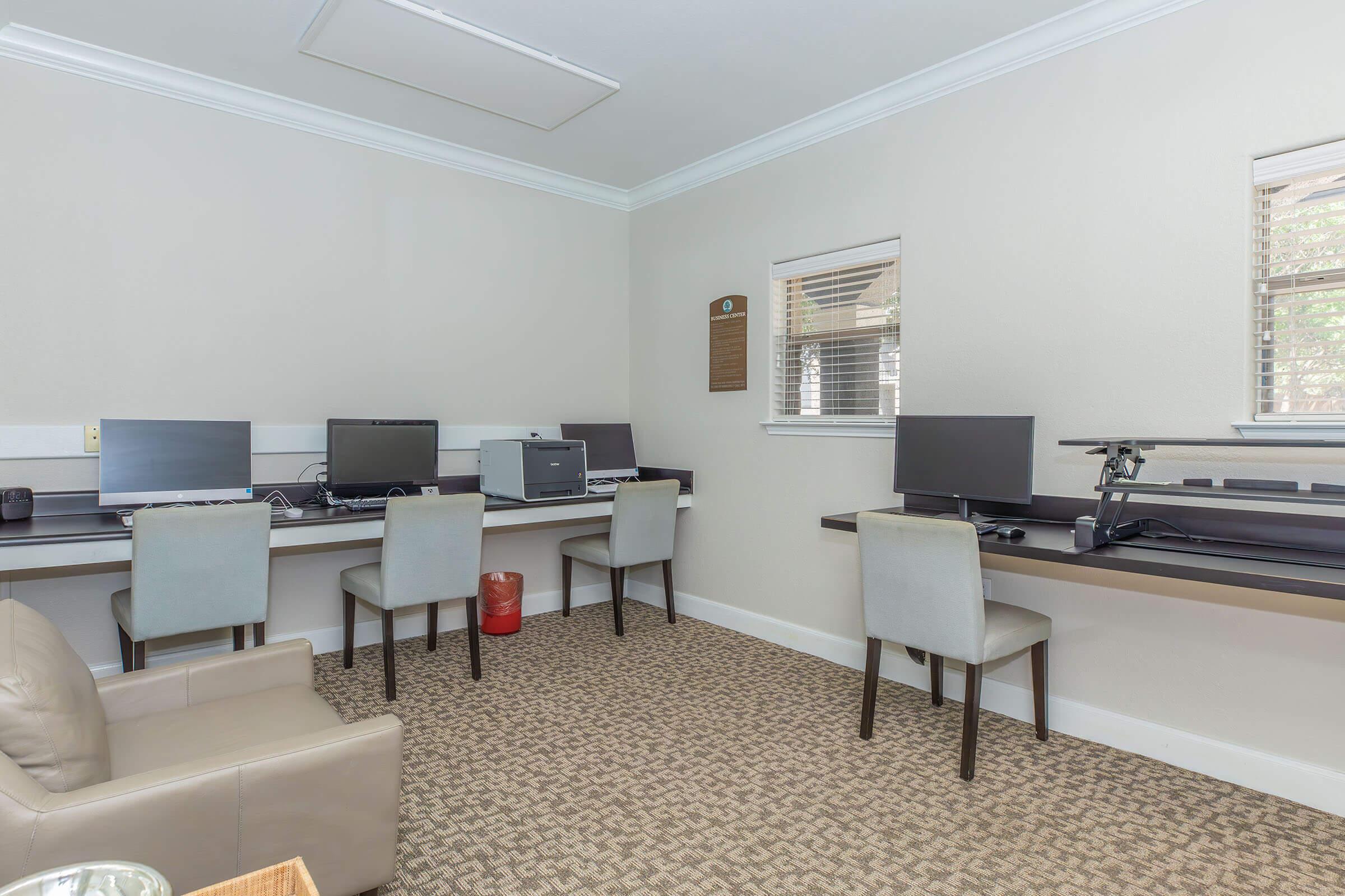 A modern office space featuring four computer stations with monitors and chairs, a grey filing cabinet, and a comfortable beige armchair in the corner. The walls are neutral-colored, and there are windows providing natural light. The floor is covered with a patterned carpet.