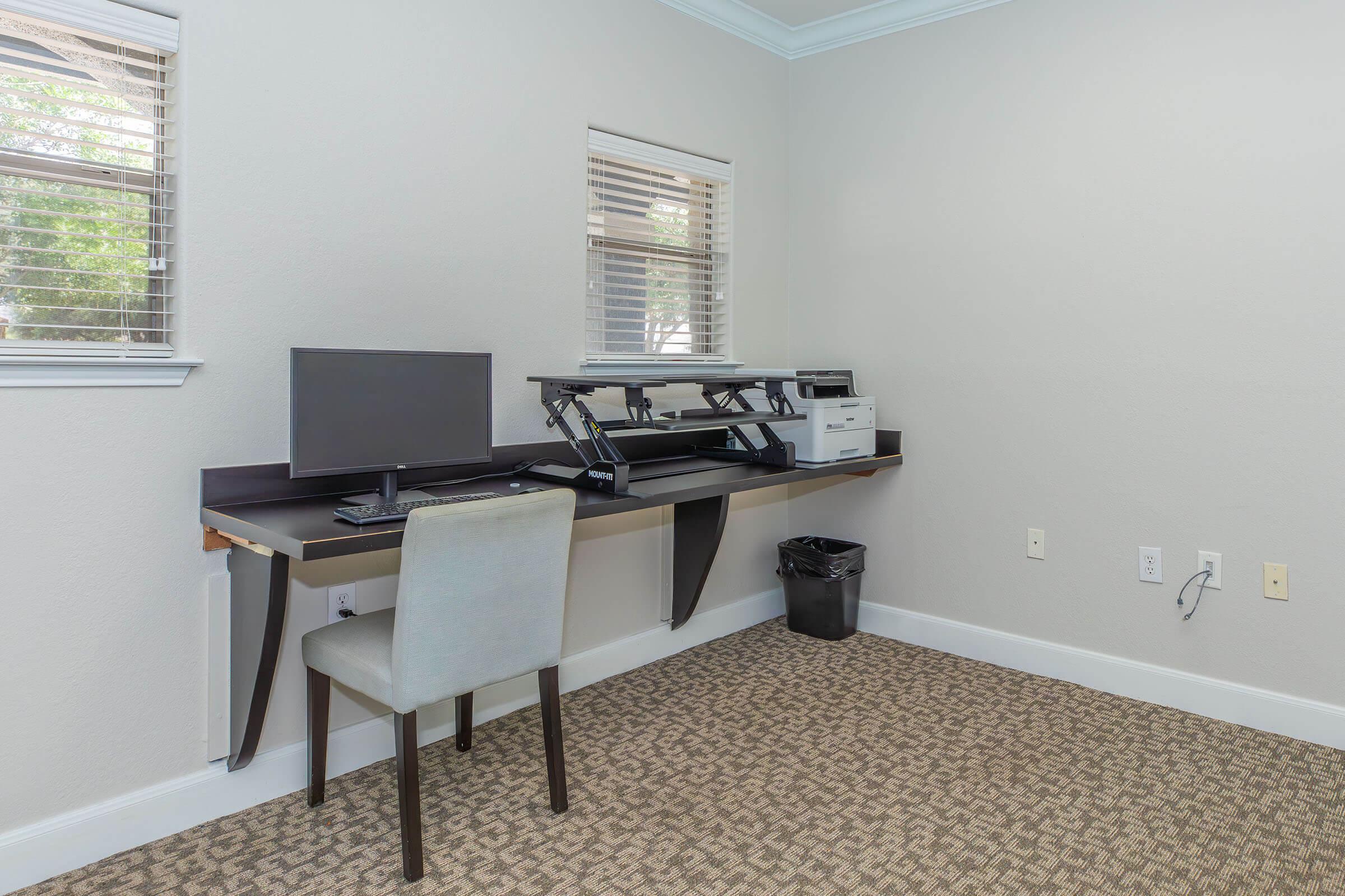 A modern, minimalistic workspace featuring a wall-mounted desk with a computer monitor, keyboard, and printer. A simple chair is placed in front of the desk. The room has light-colored walls, a carpeted floor, and two windows with blinds, allowing natural light to enter. A small trash can is positioned near the desk.