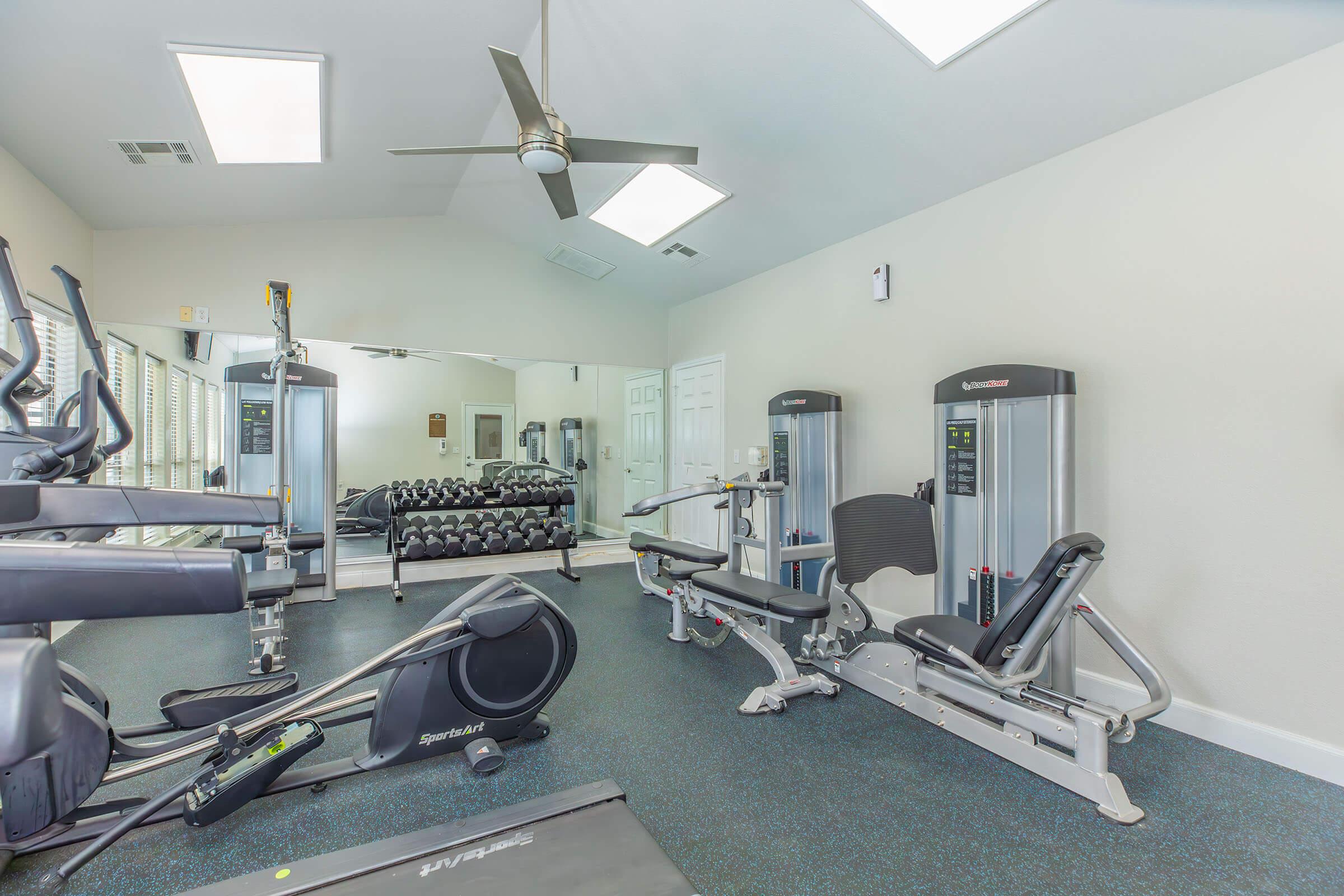 A modern gym interior with exercise equipment including a weight machine, a stationary bike, and free weights on a rack. The room features large windows for natural light, a ceiling fan, and mirrors on the wall, creating an inviting workout environment.