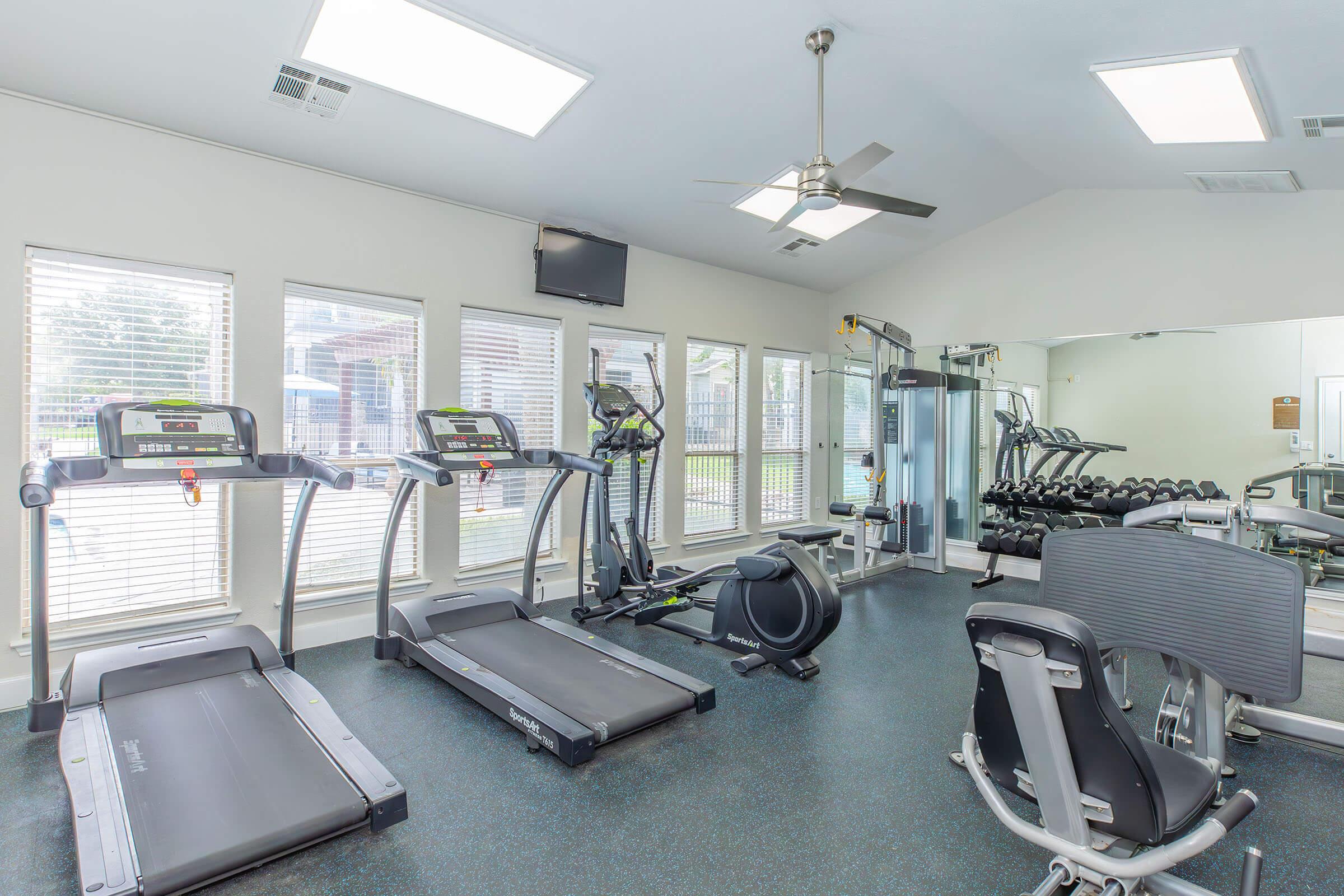 A well-lit gym interior featuring two treadmills, an exercise bike, and a weight training area with a rack of dumbbells. Large windows allow natural light to fill the space, and there is a ceiling fan for ventilation. A television is mounted on one wall, creating a motivating workout environment.