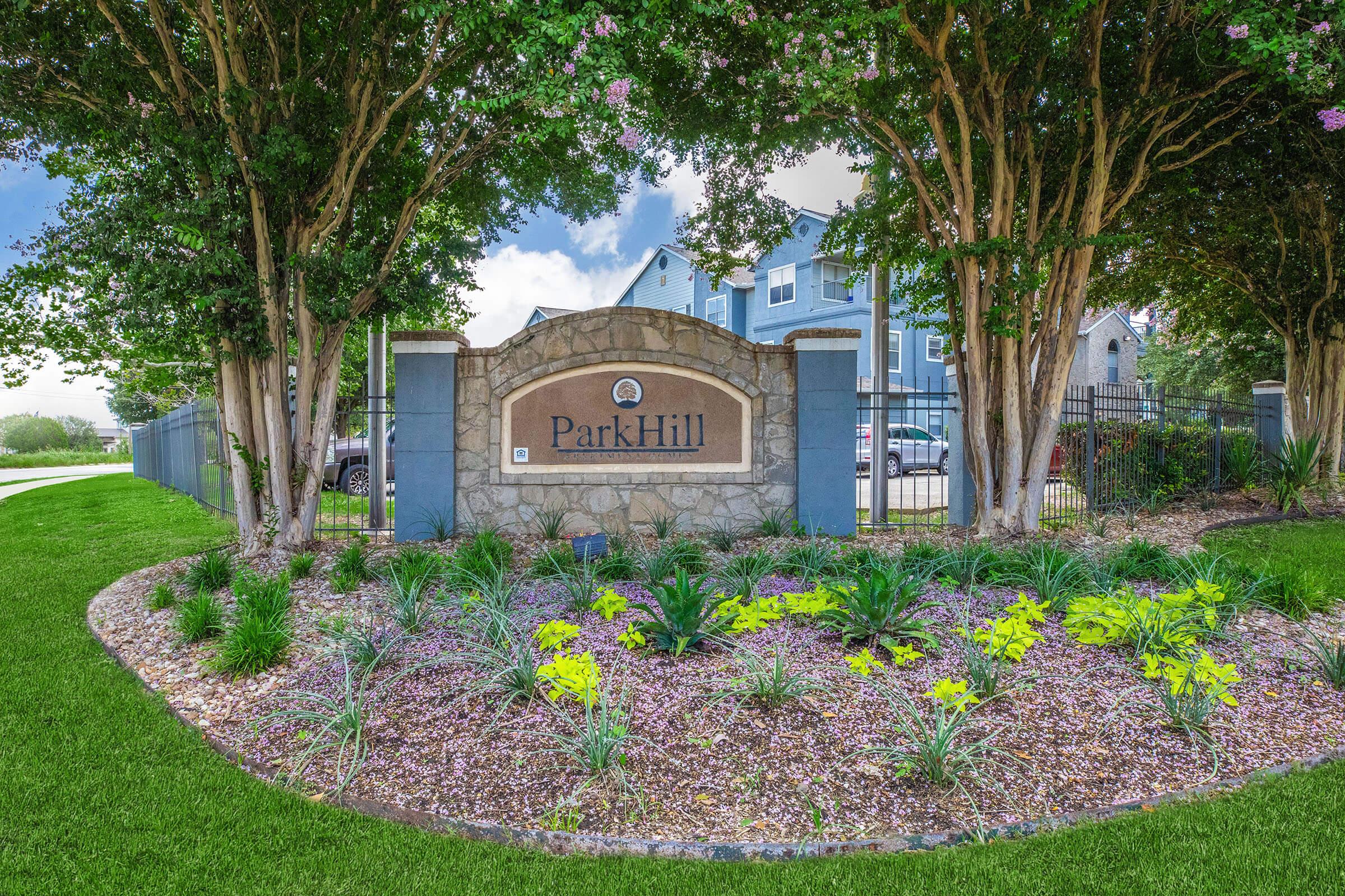 A landscaped entrance sign for "Park Hill," featuring a stone structure with the name prominently displayed. Surrounding the sign are neatly arranged flowers and plants, set against a backdrop of trees and a residential building in the distance. The scene is bright and inviting, suggesting a community space.