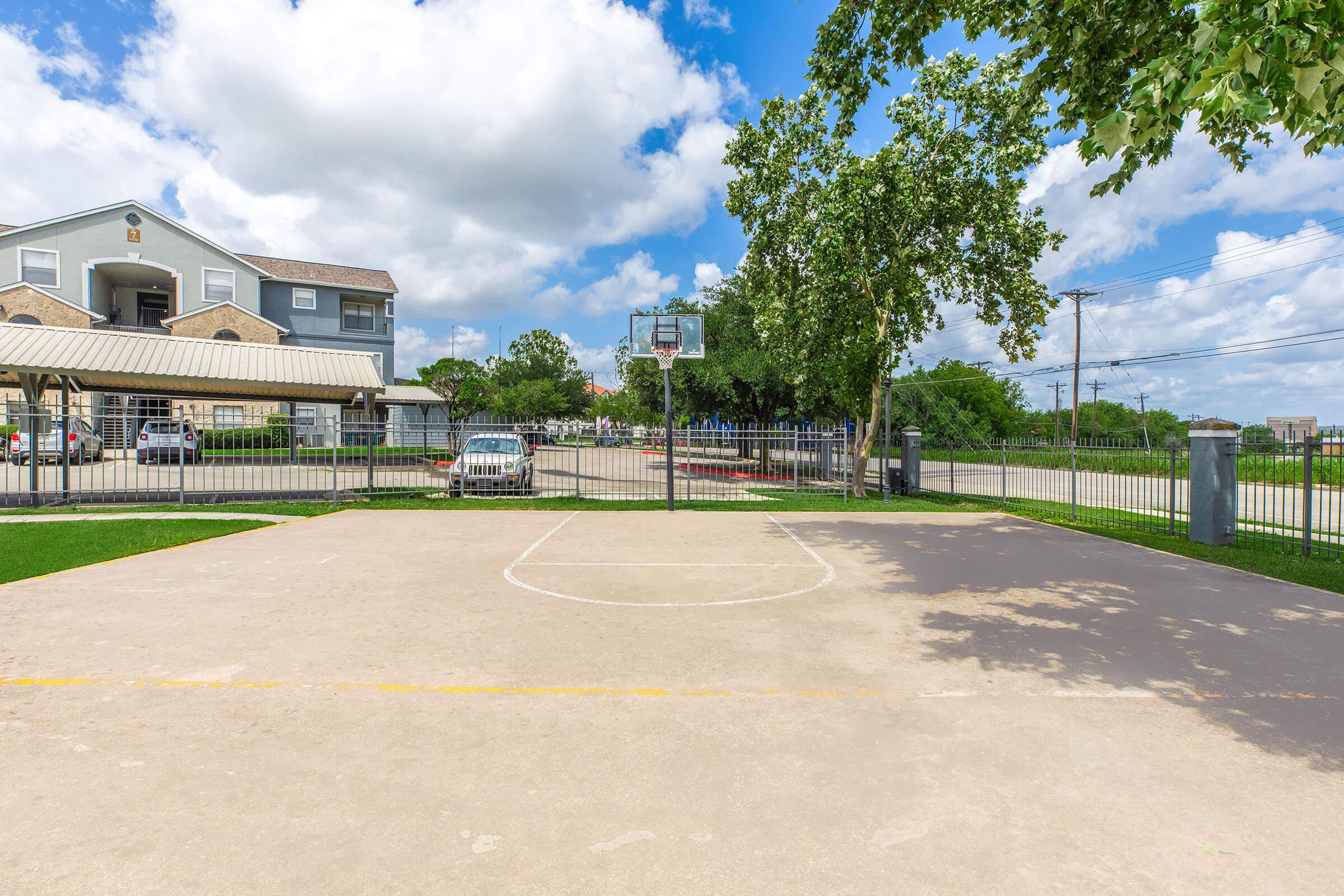 A basketball court with a hoop visible at one end. The court is surrounded by a fence and features a smooth concrete surface. In the background, a residential building and some trees are visible under a partly cloudy sky, along with a parked car and a road.