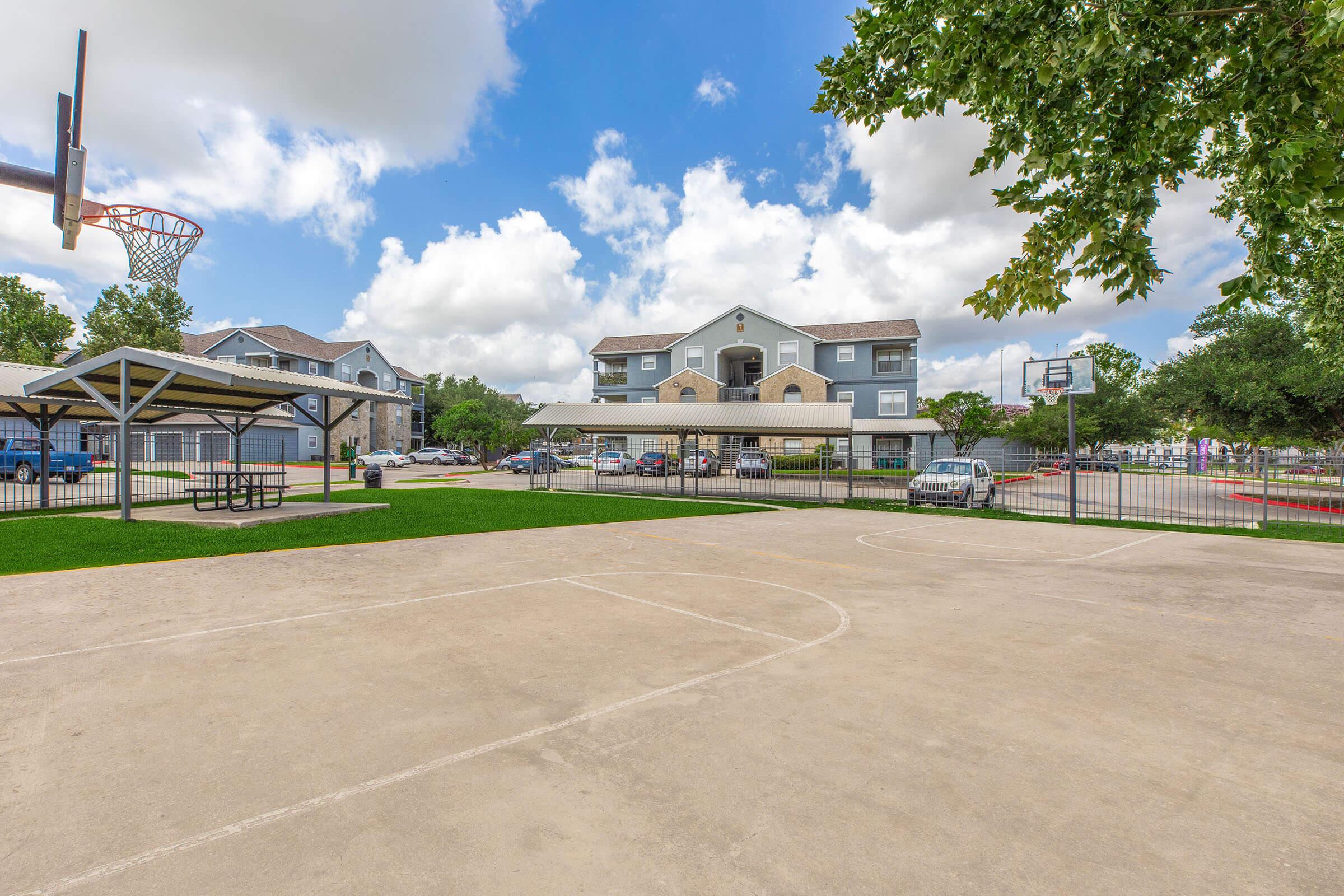 A basketball court with a hoop and a seating area under a shelter. In the background, a multi-story residential building surrounded by trees and green grass. The sky is partly cloudy, and there are parked cars nearby.