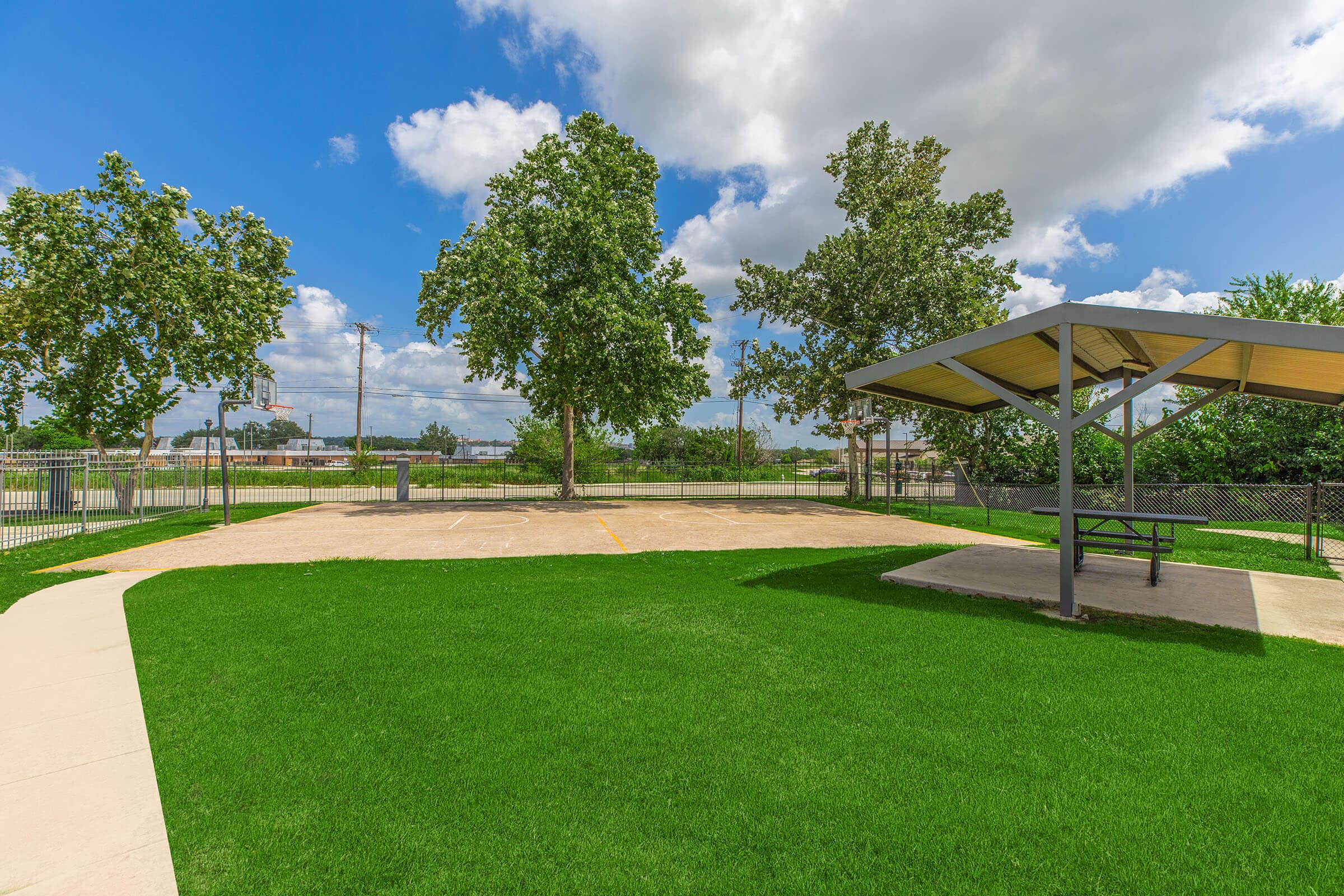 A bright outdoor area featuring a paved court surrounded by lush green grass. Tall trees provide shade, and a covered picnic area with a table is visible. The sky is partly cloudy, and there's a fence enclosing the space, with a basketball hoop in the background.