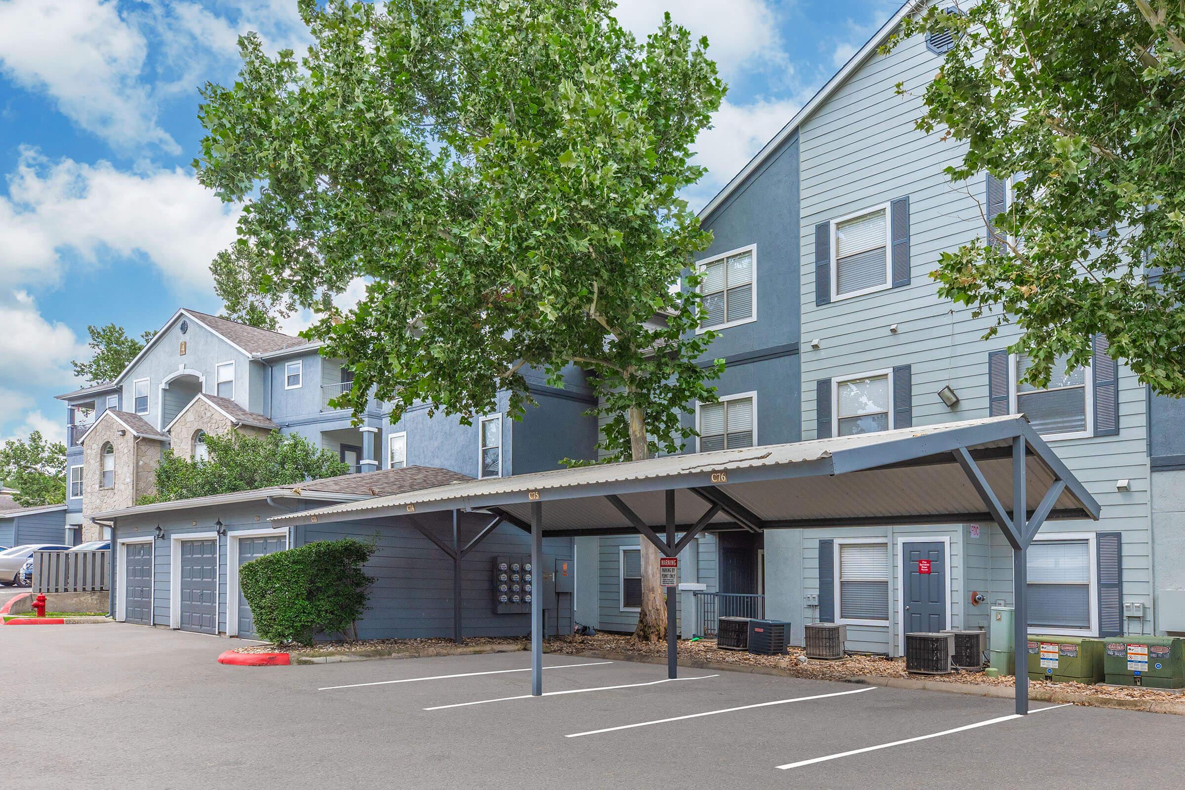 Two-story residential building with a gray exterior, featuring multiple garage doors and covered parking spaces. Surrounding landscape includes green trees and shrubs, with a clear blue sky and fluffy white clouds in the background. The area appears well-maintained and inviting.