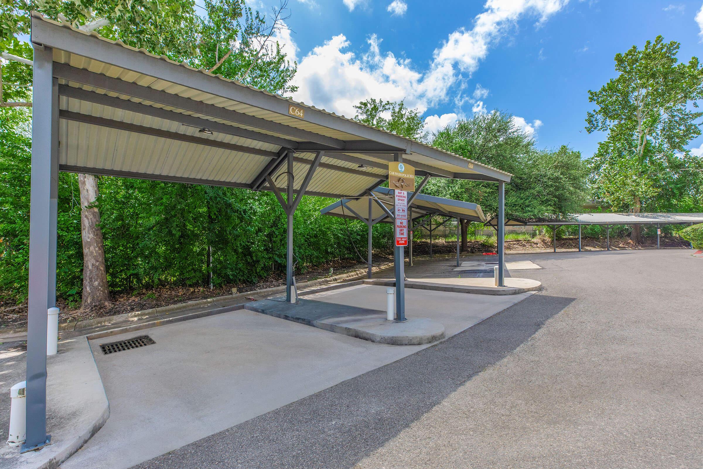 Covered parking area with several empty spaces, surrounded by trees and greenery. The sky is partly cloudy, creating a bright atmosphere. A sign stands near the entrance, indicating parking regulations or information. The pavement is clean and well-maintained.