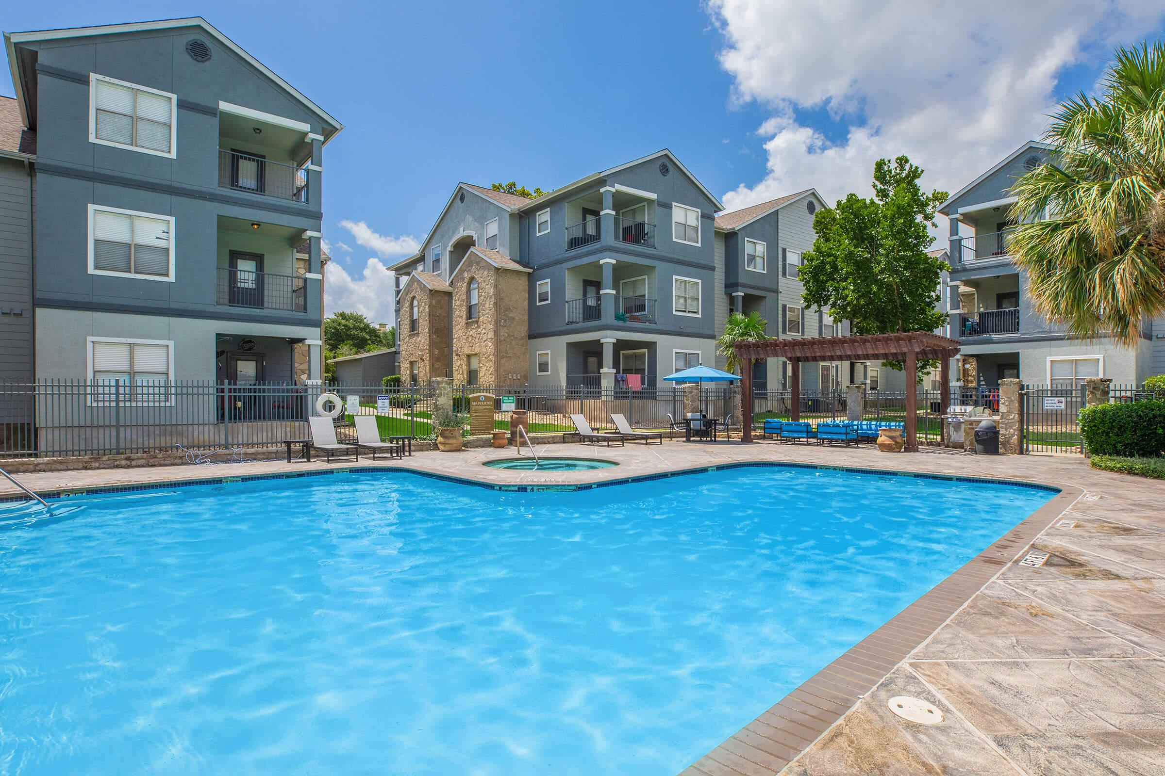 A crystal-clear swimming pool surrounded by apartment buildings. Lounge chairs are positioned around the pool, with an umbrella providing shade. Lush greenery and palm trees enhance the outdoor space, while a grill area and hot tub are visible in the background under a bright blue sky with fluffy white clouds.