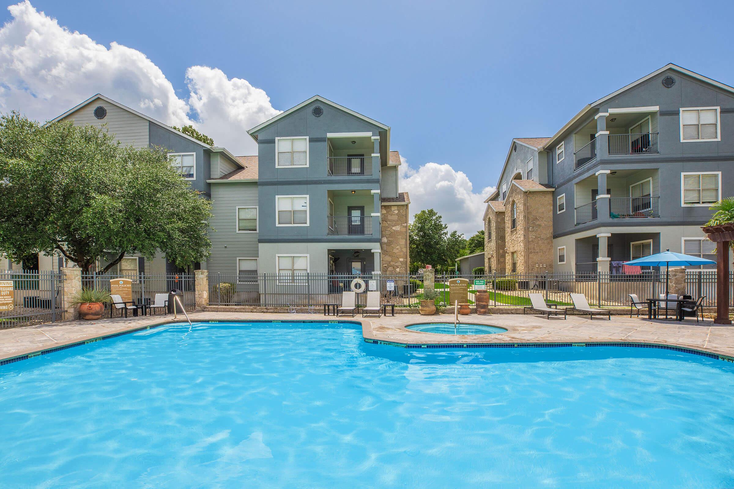 A bright blue pool surrounded by lounge chairs, with two multi-story apartment buildings in the background. Tree foliage provides shade, and a hot tub is positioned next to the pool. The sky is partly cloudy, adding to the relaxing atmosphere of the pool area.