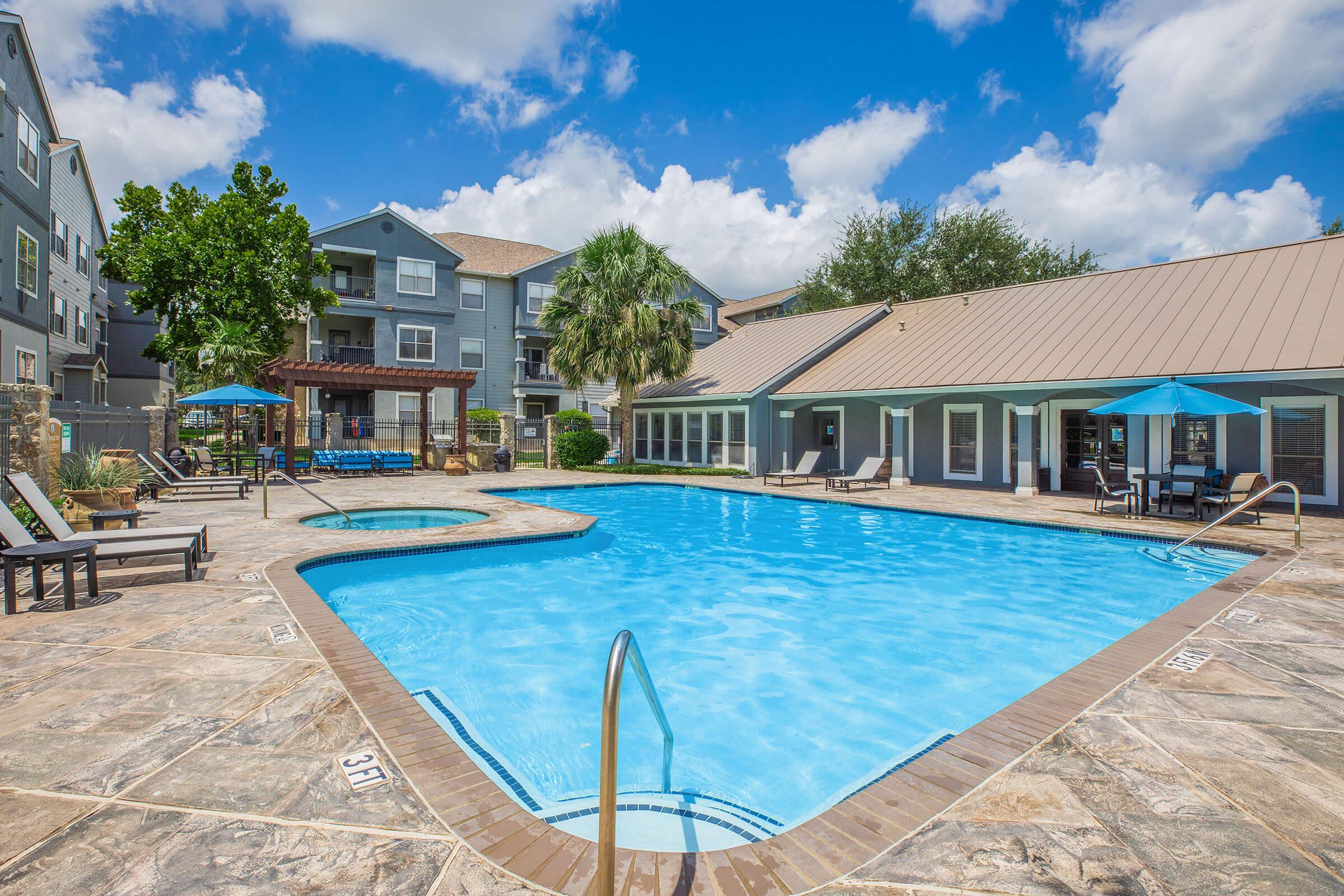 A clear blue swimming pool surrounded by lounge chairs and umbrellas, with trees and an apartment building in the background. The scene is set under a partly cloudy sky, creating a relaxing outdoor atmosphere.