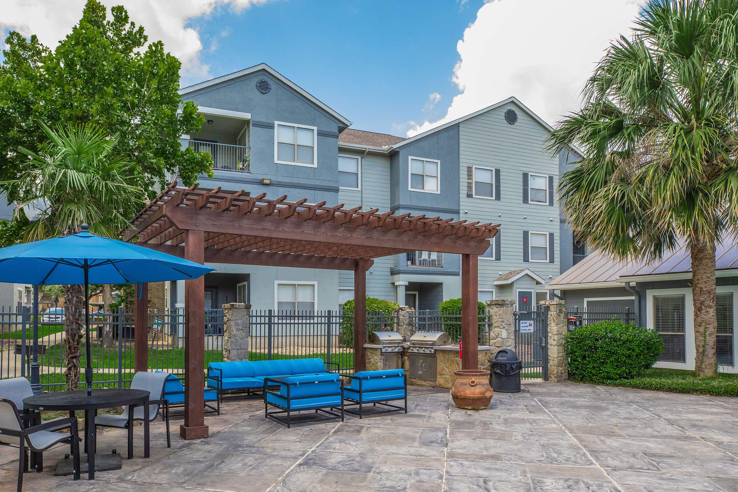 Outdoor view of an apartment community featuring a pergola with blue seating and a round table. There is a green lawn, decorative palm trees, and a fence surrounding the area. The complex is two stories, with a gray and blue exterior and multiple windows. A barbecue grill and trash can are visible nearby.