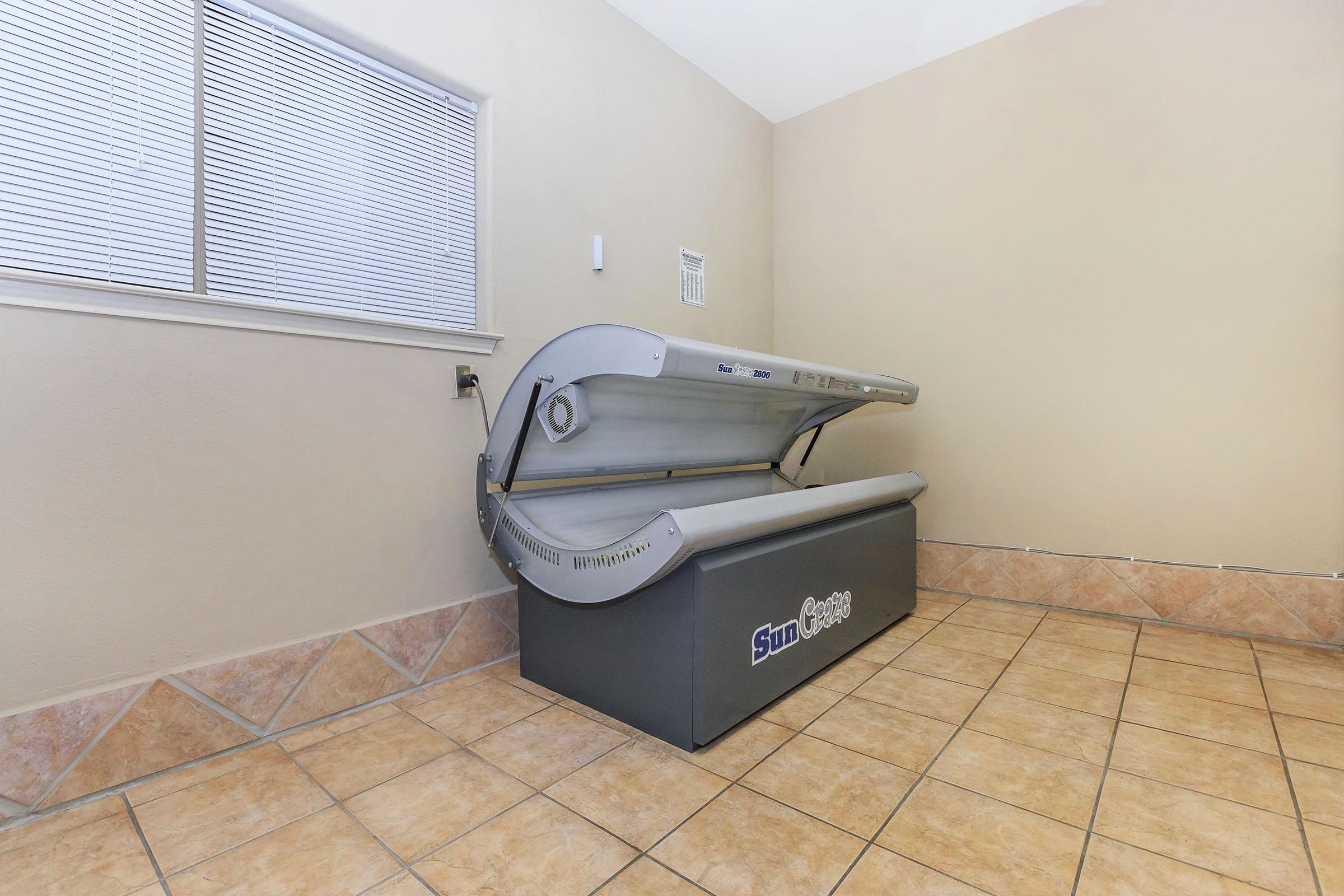 A sun tanning bed is positioned in a well-lit room with beige walls and tiled flooring. The tanning bed is closed and branded with the name "Sun Capsule." A window with blinds allows natural light into the space, contributing to a bright and neutral environment.