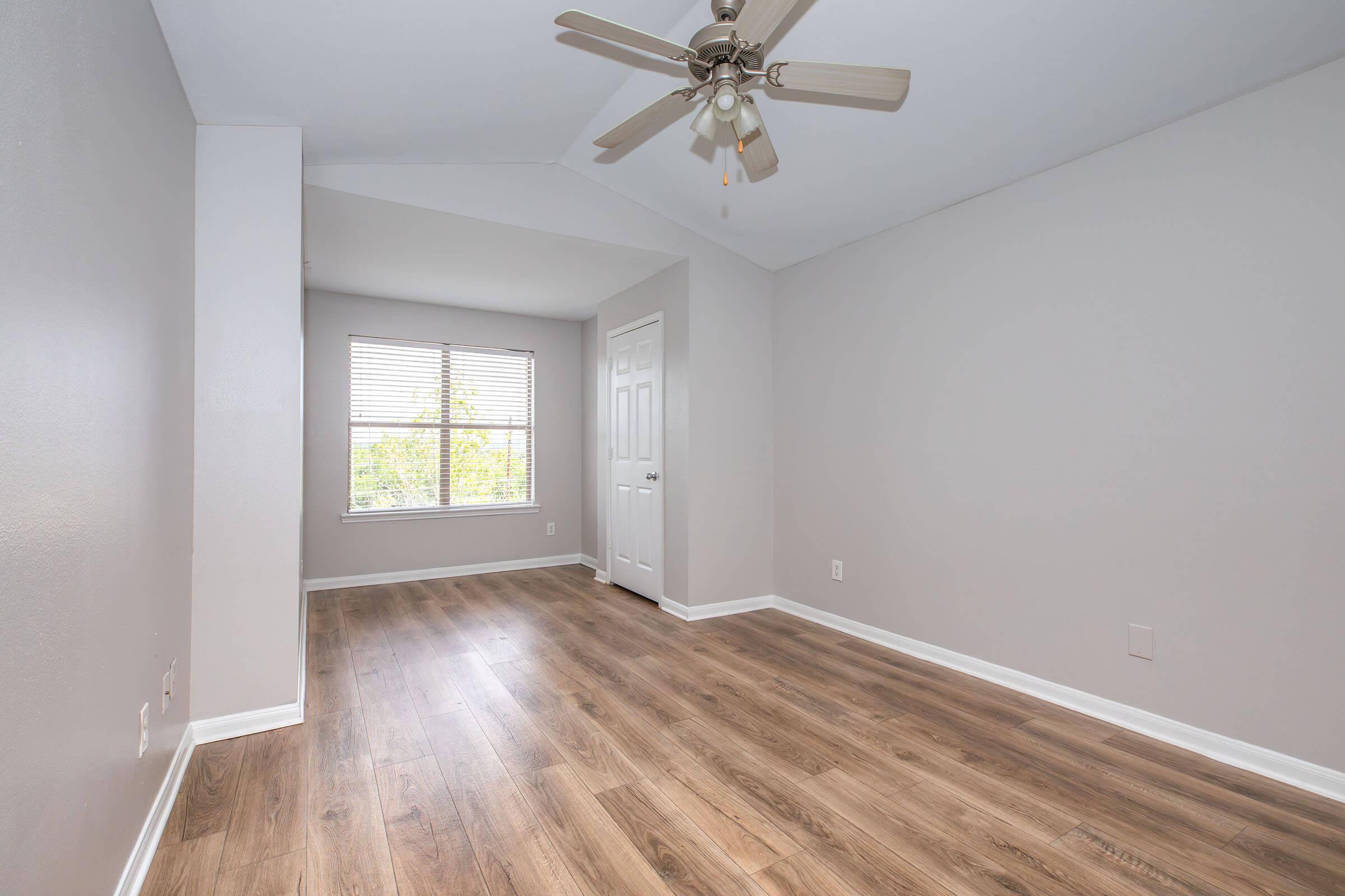 A spacious, well-lit room with light gray walls and hardwood flooring. A ceiling fan hangs from the sloped ceiling, and a window with blinds allows natural light. A door leads to another area, creating an inviting and airy atmosphere.