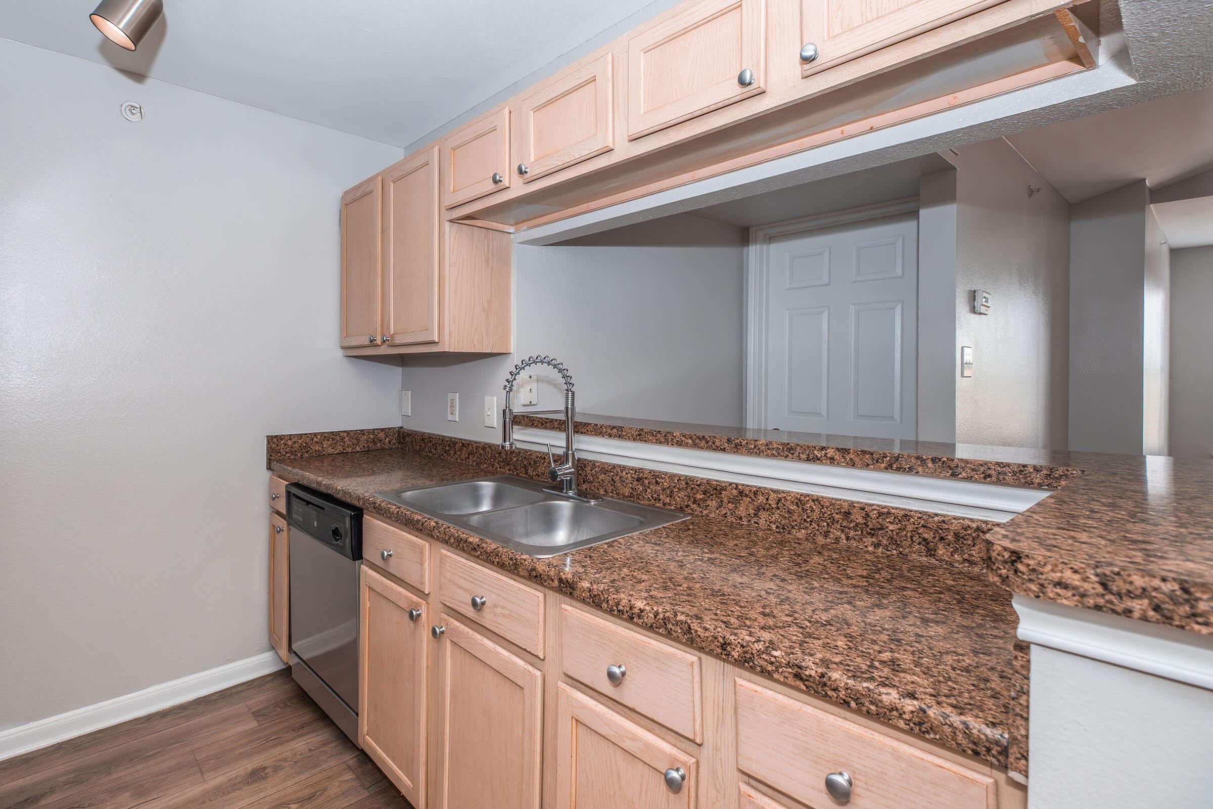 Modern kitchen with light wooden cabinets, granite countertops, and stainless steel appliances. The sink is positioned between the cabinets, and there is a dishwasher next to it. A mirror reflects the room, providing a sense of space. The flooring is a light wood laminate.