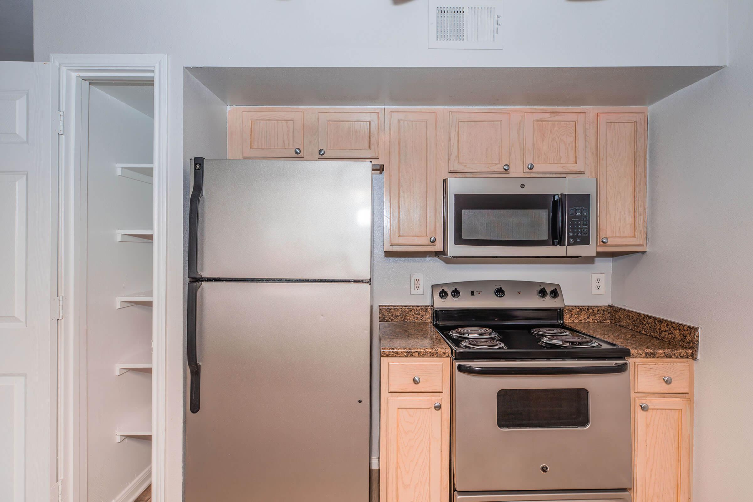 A modern kitchen featuring a stainless steel refrigerator, a microwave above a black stove with two burners, and light wooden cabinetry. The countertop is made of dark granite. A pantry door is visible on the left, and the wall is painted light gray.