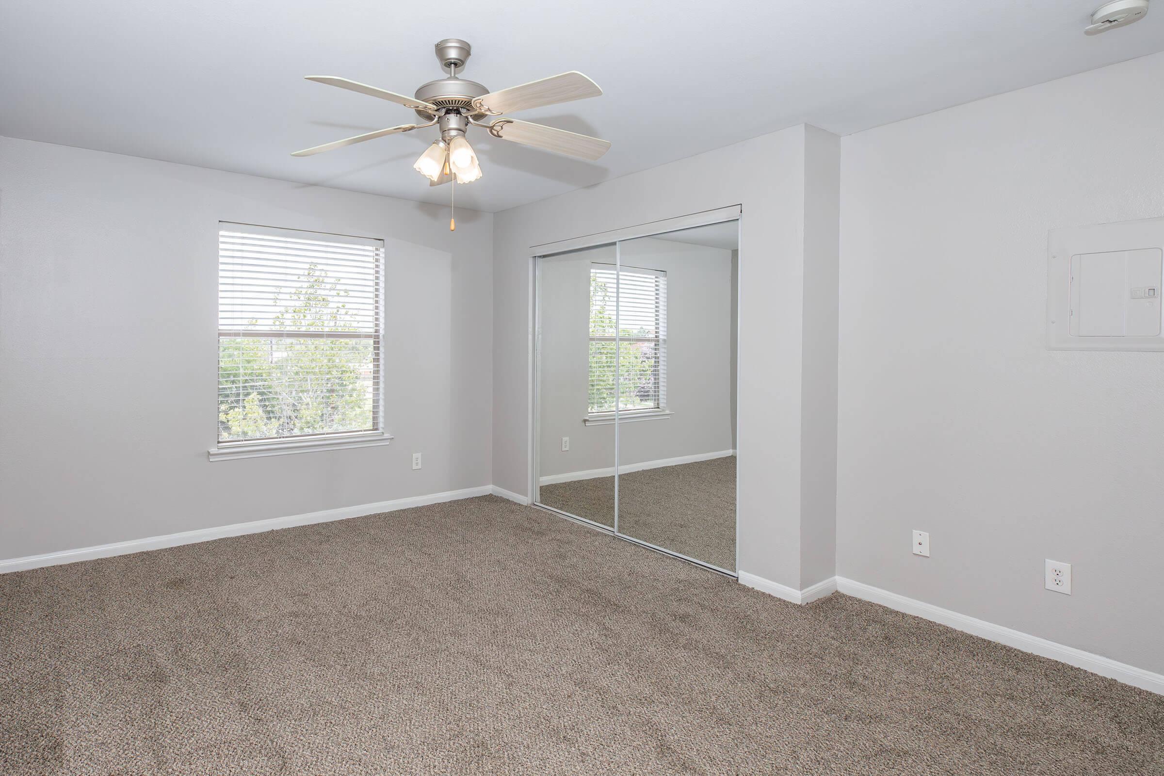 A spacious, neutral-toned bedroom featuring light-colored walls, a ceiling fan with three lights, and large windows with blinds. The floor is covered in plush carpet, and a mirrored closet with sliding doors is visible on one side. Natural light enhances the room's airy ambiance.