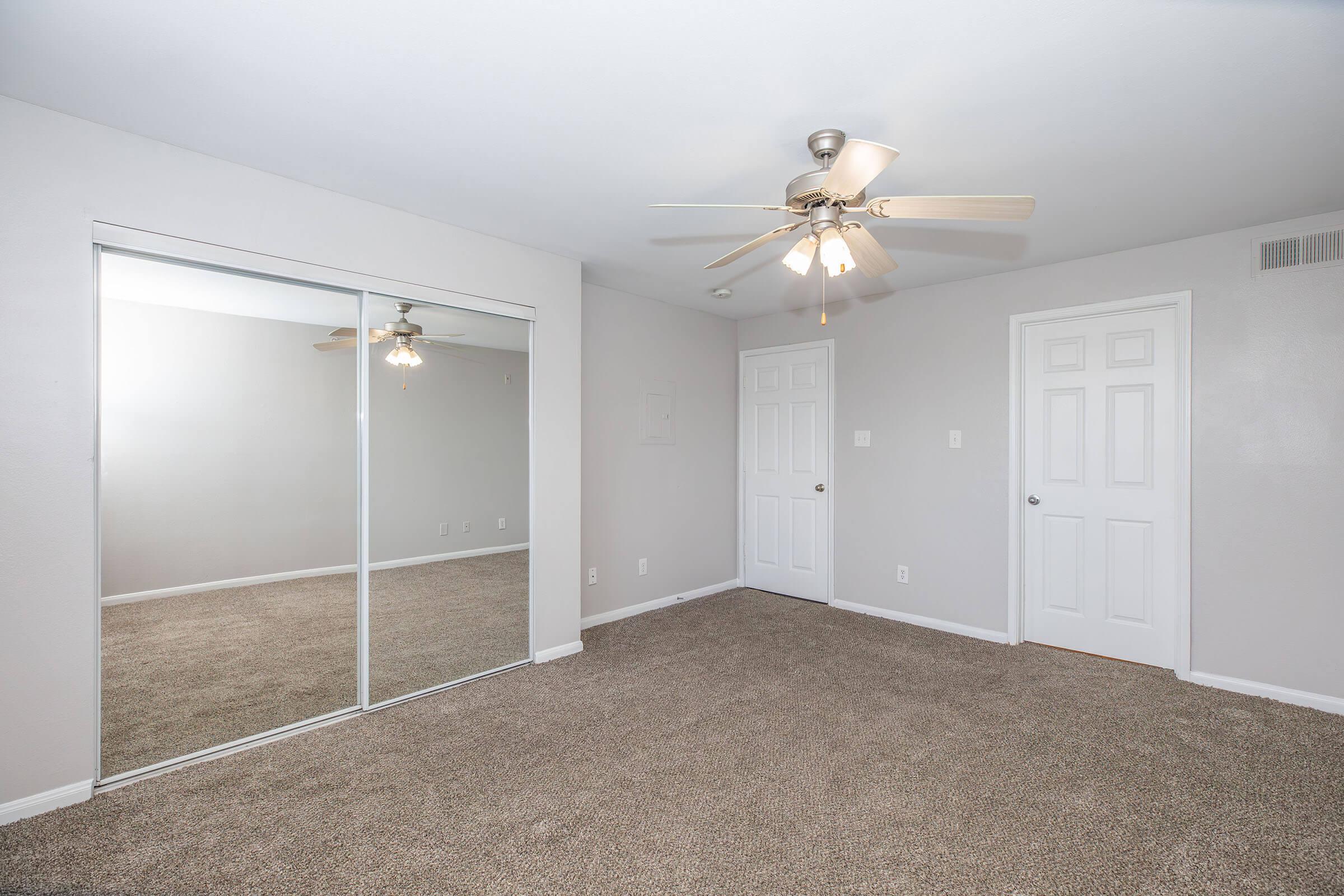 A spacious, well-lit bedroom featuring light gray walls and beige carpet. It includes a ceiling fan with light, a mirrored closet door on one wall, and two white doors leading to other rooms. The space is tidy and empty, ready for furniture.