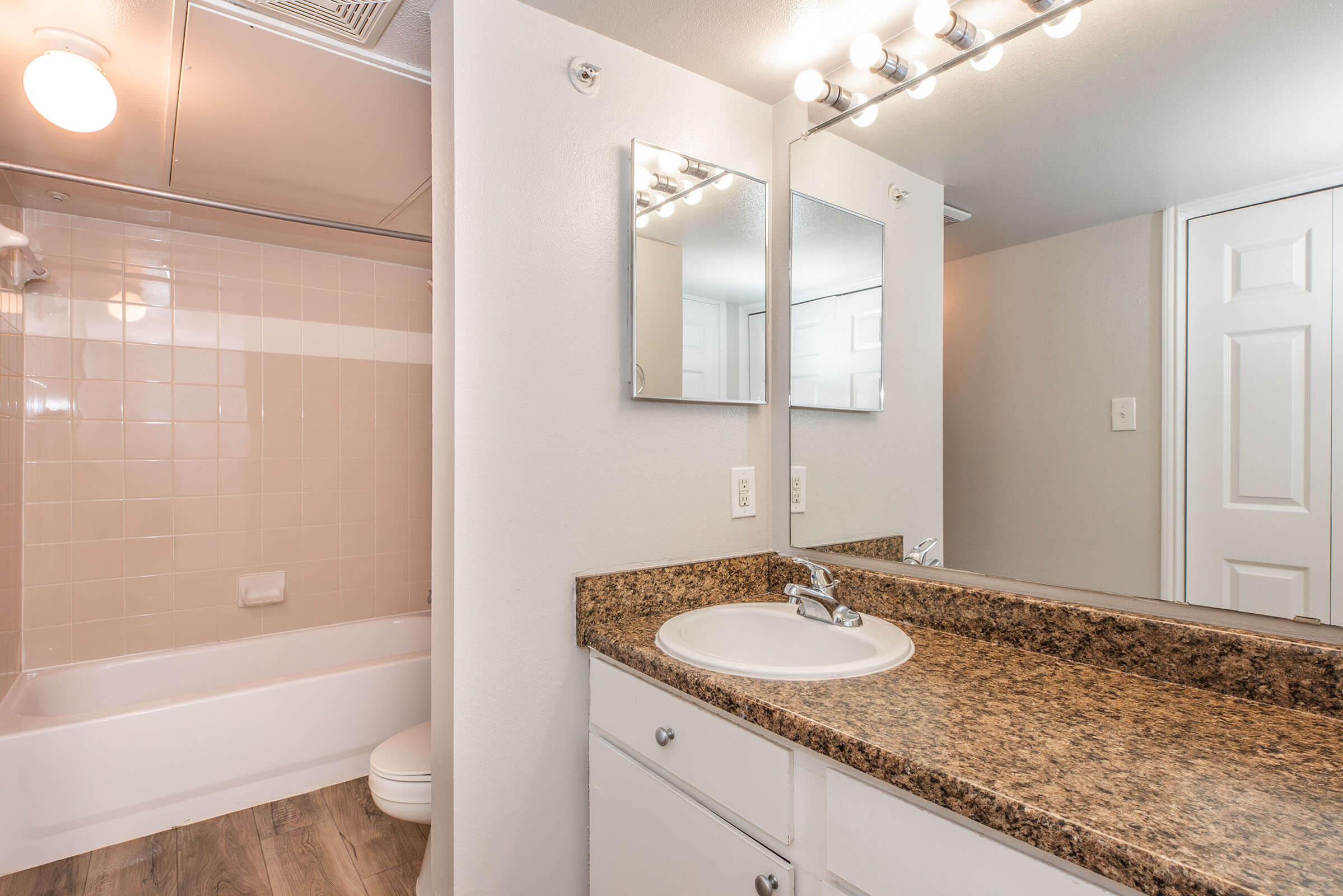 A modern bathroom featuring a granite countertop with a sink, a large mirror, and bright vanity lights. The area includes a shower-tub combo with light-colored tiles, white cabinetry, and a neutral wall color, offering a clean and spacious appearance. A door leads to an adjacent room.