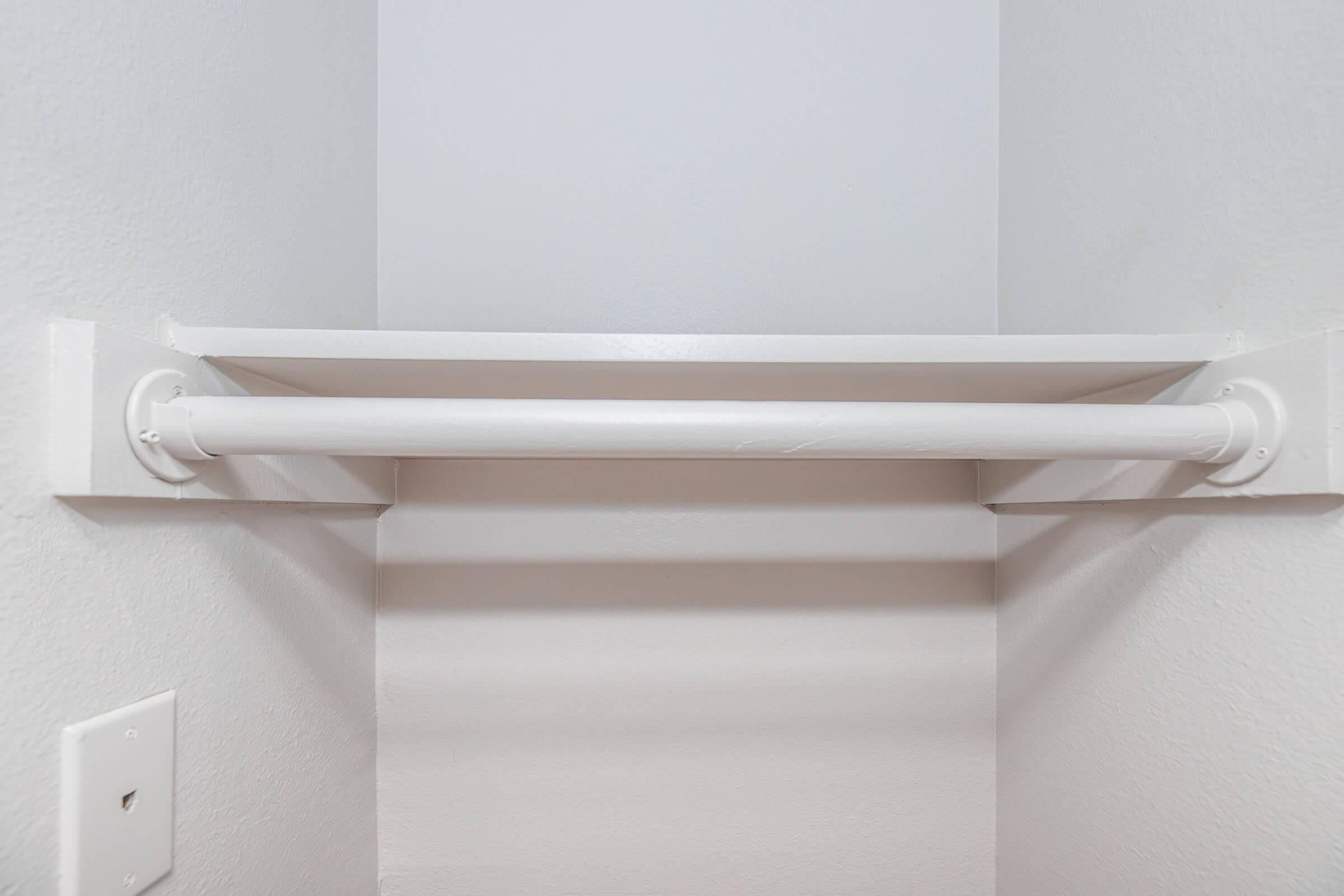 A simple white closet space featuring a horizontal rod mounted on the wall for hanging clothes, with a light gray wall in the background and an electrical outlet visible below the rod. The overall look is clean and minimalistic.