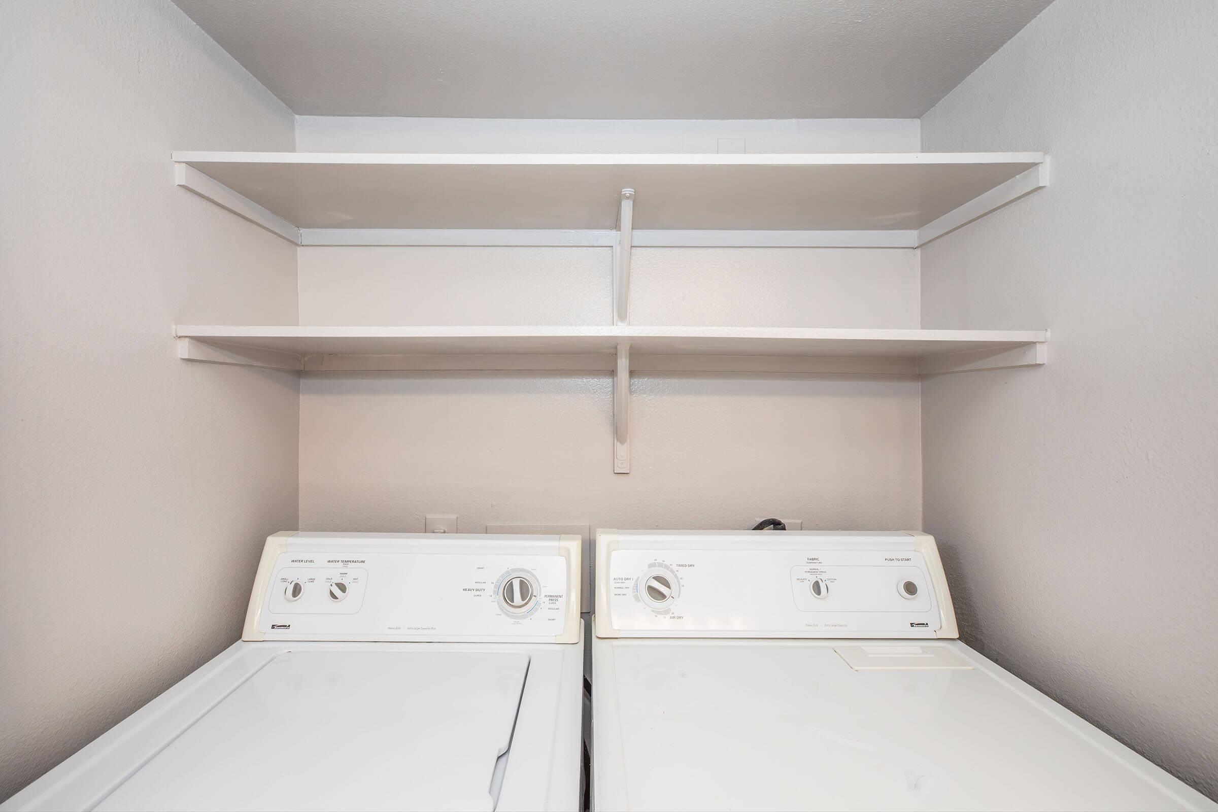 A clean laundry space featuring a white washer and dryer side by side, with two white shelves above them. The walls are painted light gray, creating a neat and organized look. The shelves are empty, providing ample space for storing laundry supplies or other items.