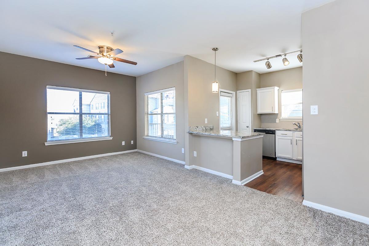 Spacious living area with light gray carpet, ceiling fan, and large windows providing natural light. To the right, a modern kitchen features white cabinets, stainless steel appliances, and a peninsula with seating. The open layout creates a welcoming atmosphere, ideal for relaxation and entertaining.