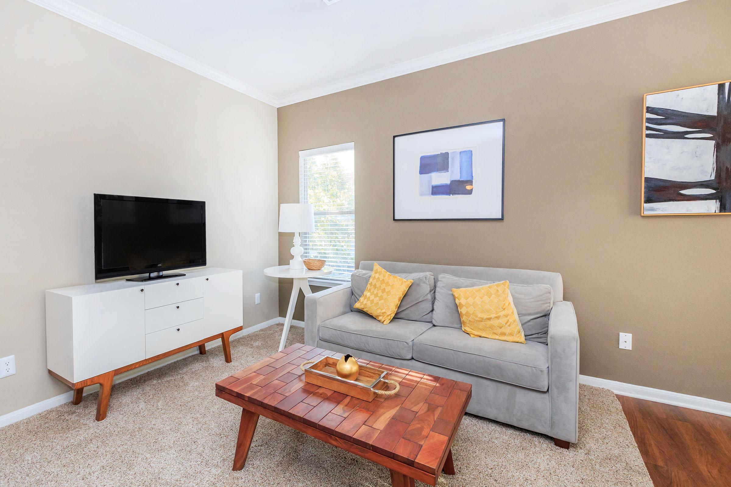 A modern living room featuring a light gray couch with yellow accent pillows, a wooden coffee table with a decorative item, a white side table with a lamp, and a television on a stylish media console. The walls are painted a warm beige, complemented by a framed artwork and natural light from a window.