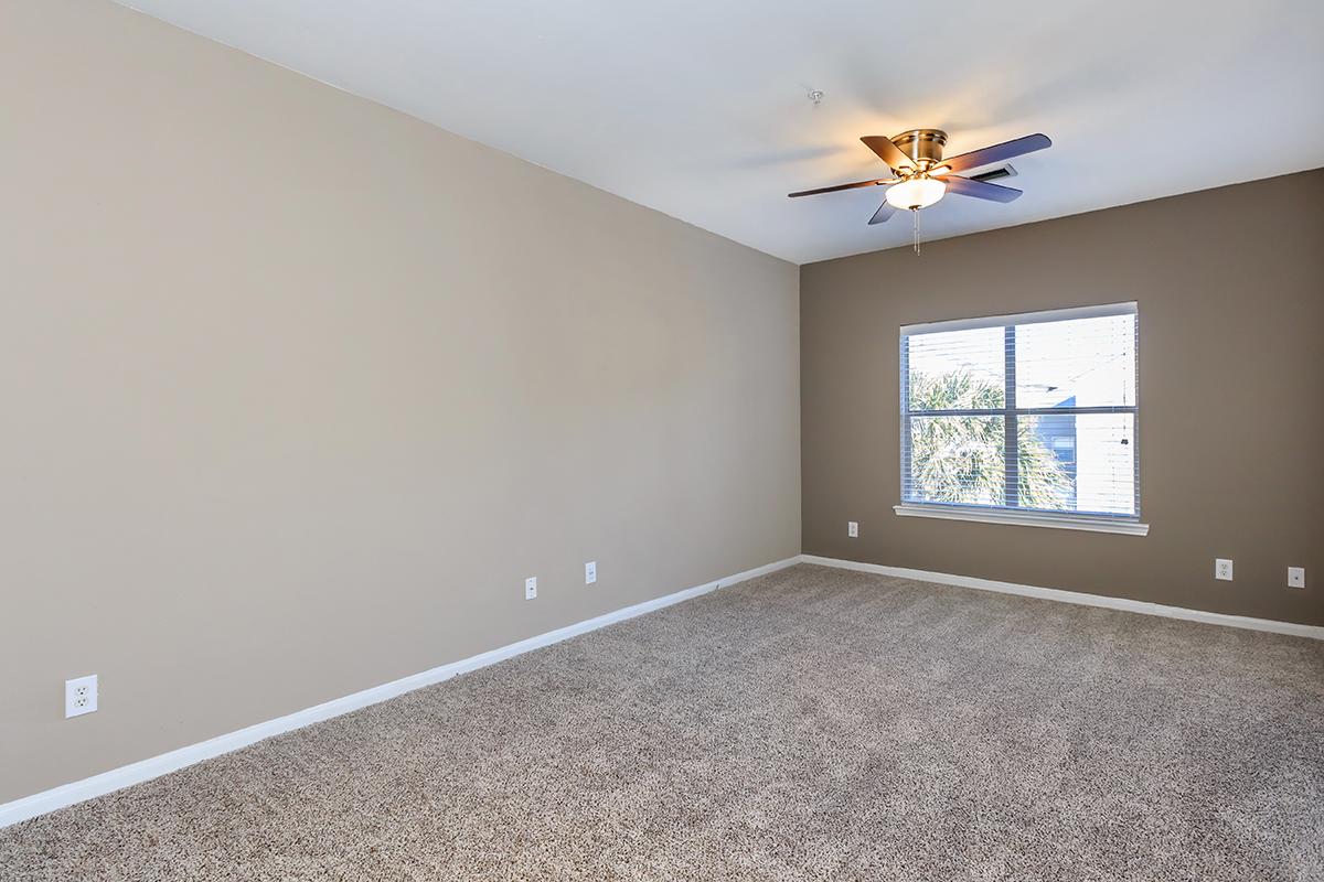 A spacious, empty room with light brown walls and a ceiling fan. The floor is covered in beige carpet, and there is a window on one side letting in natural light, showcasing a view of trees outside. The room is designed for versatility, suitable for various uses.