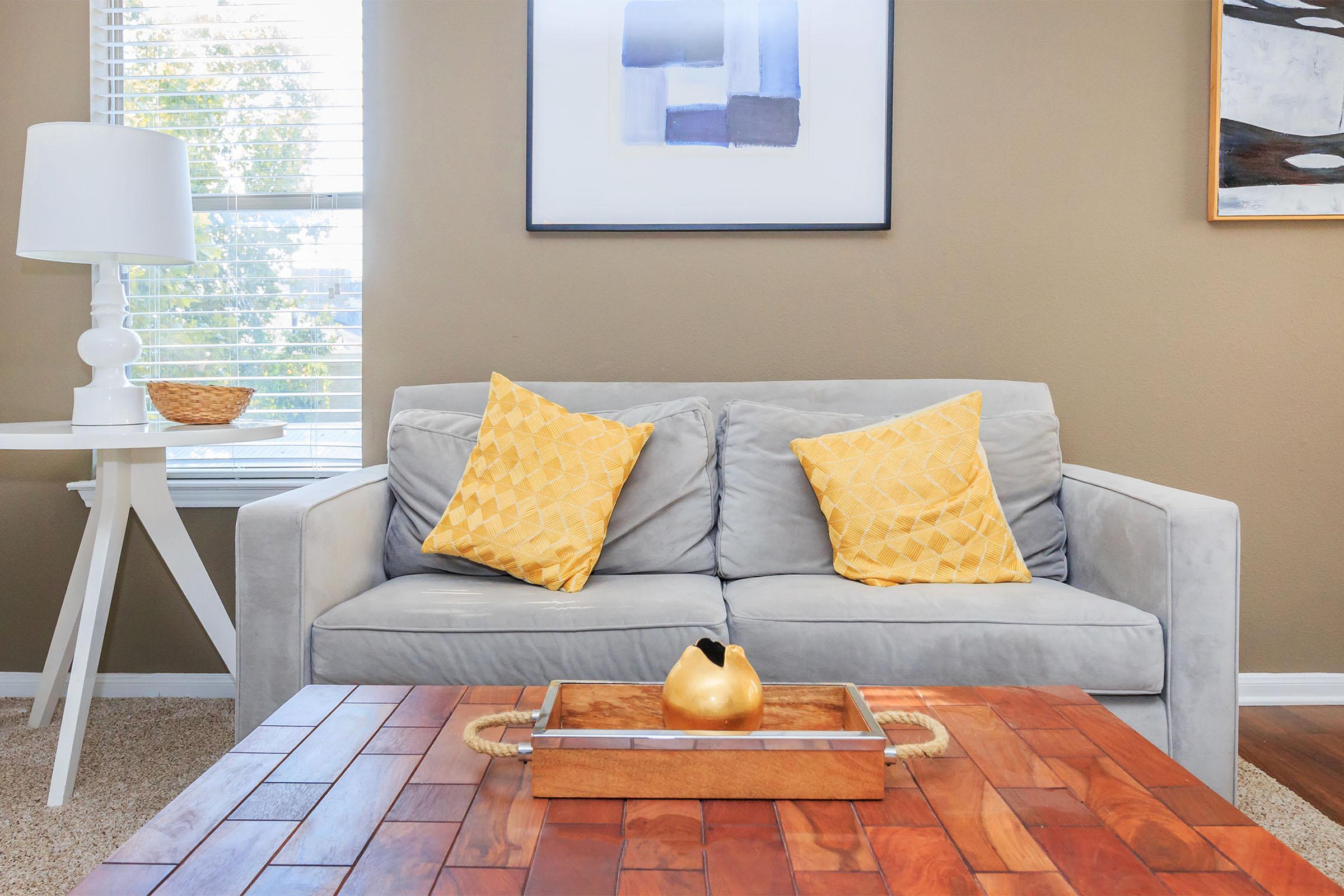 A cozy living room featuring a light gray sofa with two yellow decorative pillows. In front of the sofa is a wooden coffee table with a tray holding a decorative golden pear. A stylish lamp and a woven basket are on a side table nearby, and sunlight filters through the window, illuminating the space.