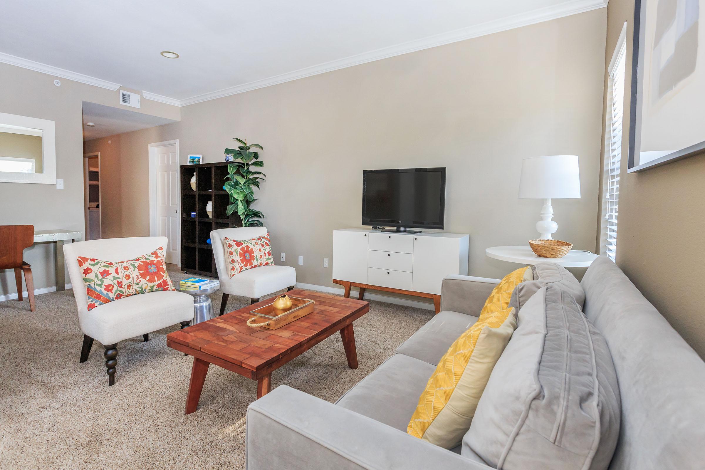 A modern living room featuring a comfortable gray sofa with decorative pillows, two white armchairs with floral patterns, a wooden coffee table with decorative items, and a white TV console. The room has light beige walls, a lamp, and a small bookshelf, creating a cozy and inviting atmosphere.