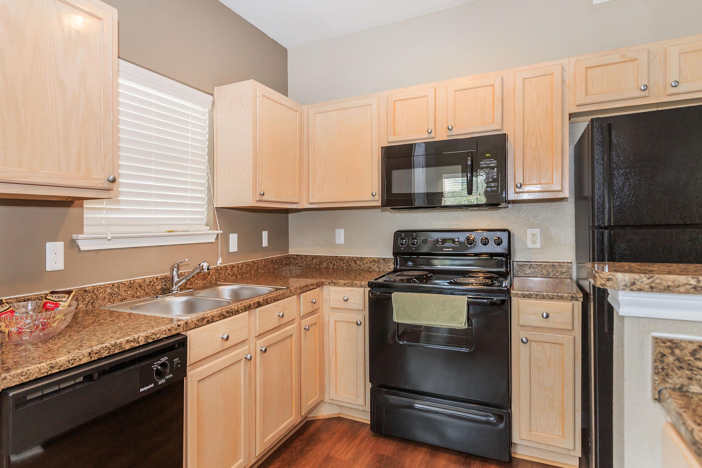 A modern kitchen featuring light wood cabinetry, a black gas stove and microwave, a double sink, and a dishwasher. The countertop is made of dark granite, and there are blinds on the window allowing natural light. The flooring is dark wood, creating a warm and inviting atmosphere.