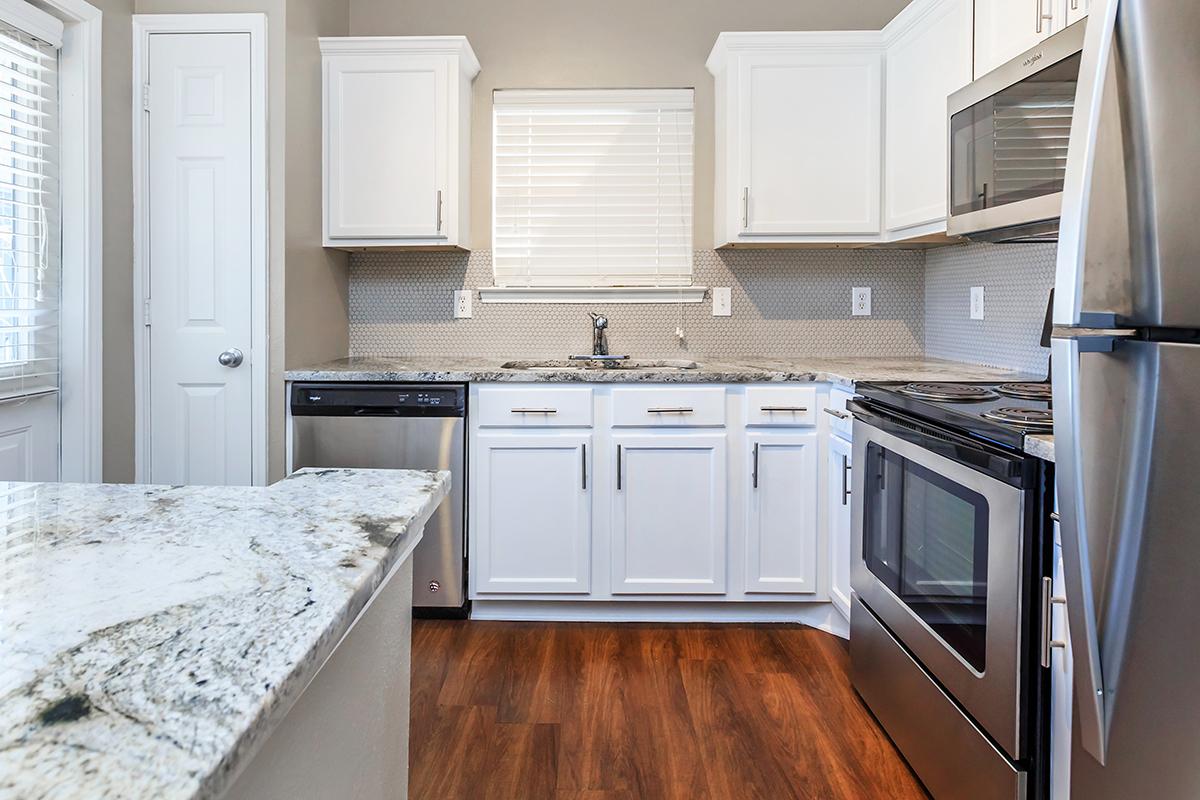 Modern kitchen featuring white cabinetry, stainless steel appliances, a granite countertop, and dark wood flooring. Natural light comes in through the window with blinds. The kitchen has a dishwasher, oven, and a sleek sink, creating a functional and stylish cooking space.