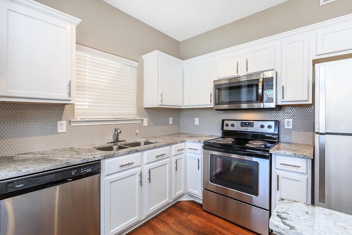 Bright, modern kitchen featuring white cabinets, stainless steel appliances including a microwave and oven, a sink with a window above, and granite countertops. The floor is made of rich wood, and there is a patterned backsplash made of small tiles.