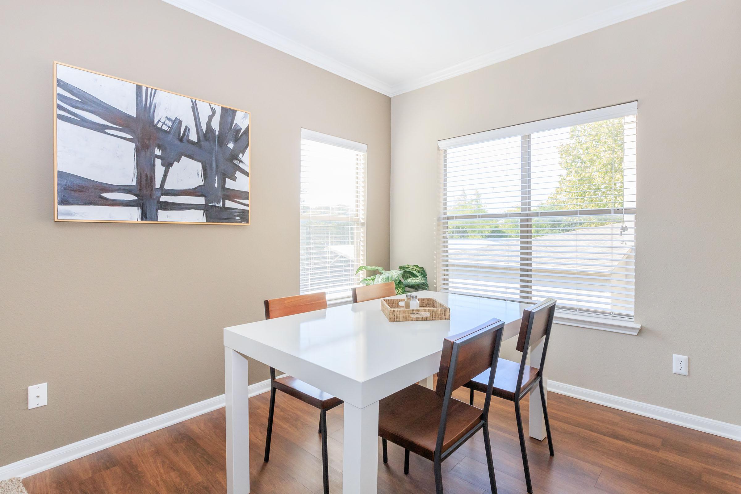 A bright dining area featuring a white table surrounded by four wooden chairs. The walls are painted in a light beige color, and there's a large abstract artwork hanging on the wall. A window with blinds lets in natural light, and a small plant is positioned on the table.